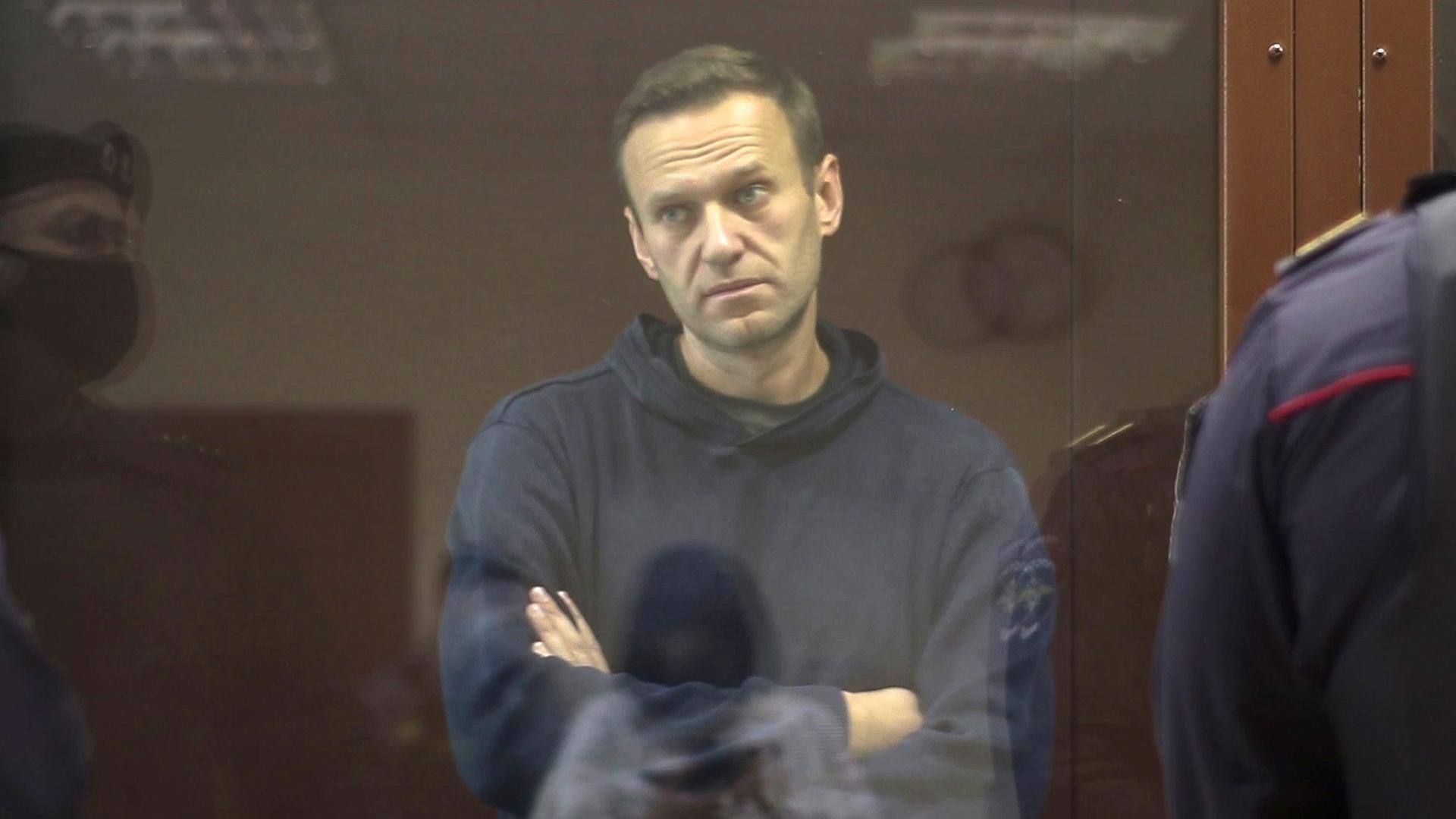 Alexei Navalny is shown wearing a blue hooded sweatshirt and standing with his arms folded in a glass cage in a Russian courtroom.