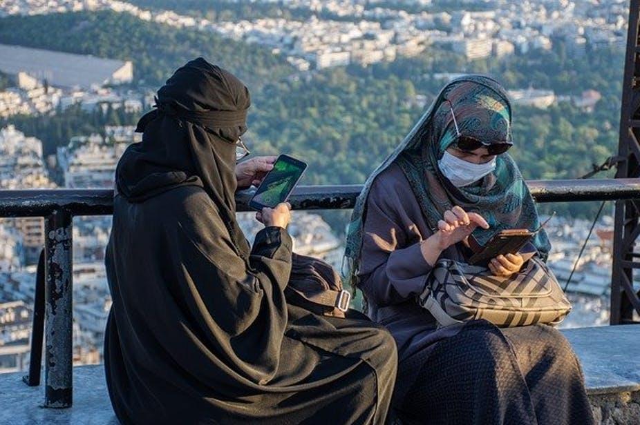 Two women sit outside, using their cell phones. They are wearing face masks in addition to head coverings. 