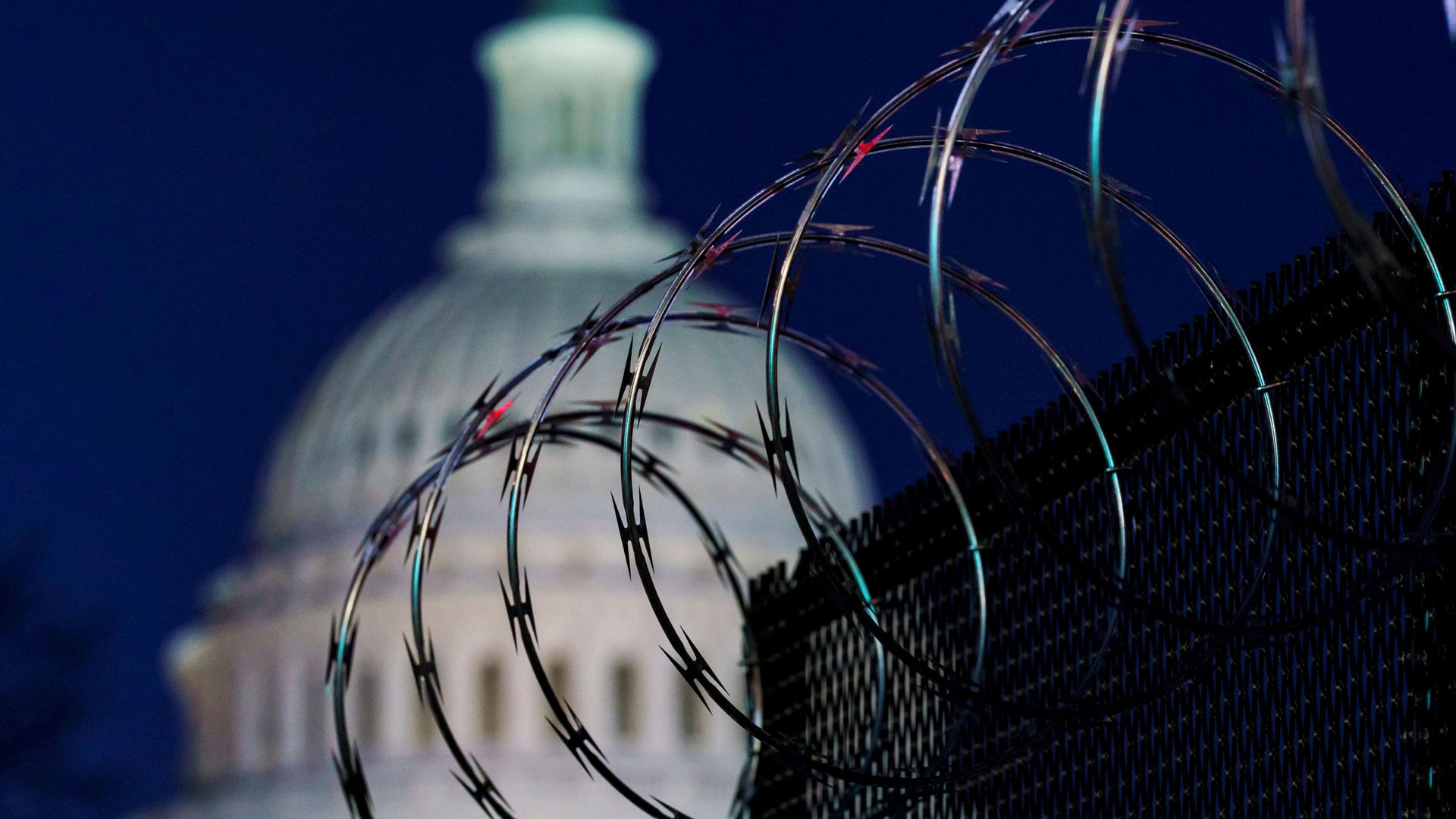 A fence with large rings of barbed wire is shown in the nearground with the dome of the US Capitol is soft focus in the background.