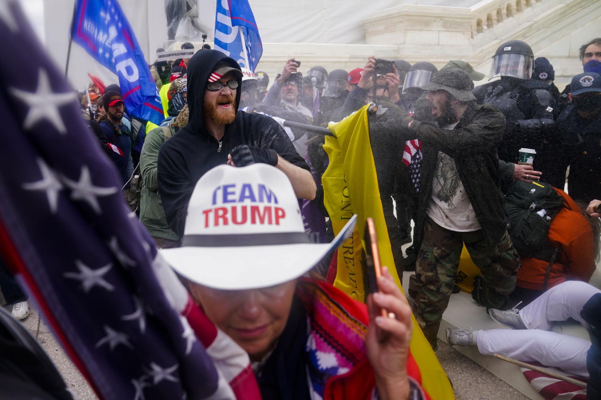 Several people wearing pro-Trump paraphernalia are shown outside of the US Capitol building with police wearing helmet and face guards shown opposite.