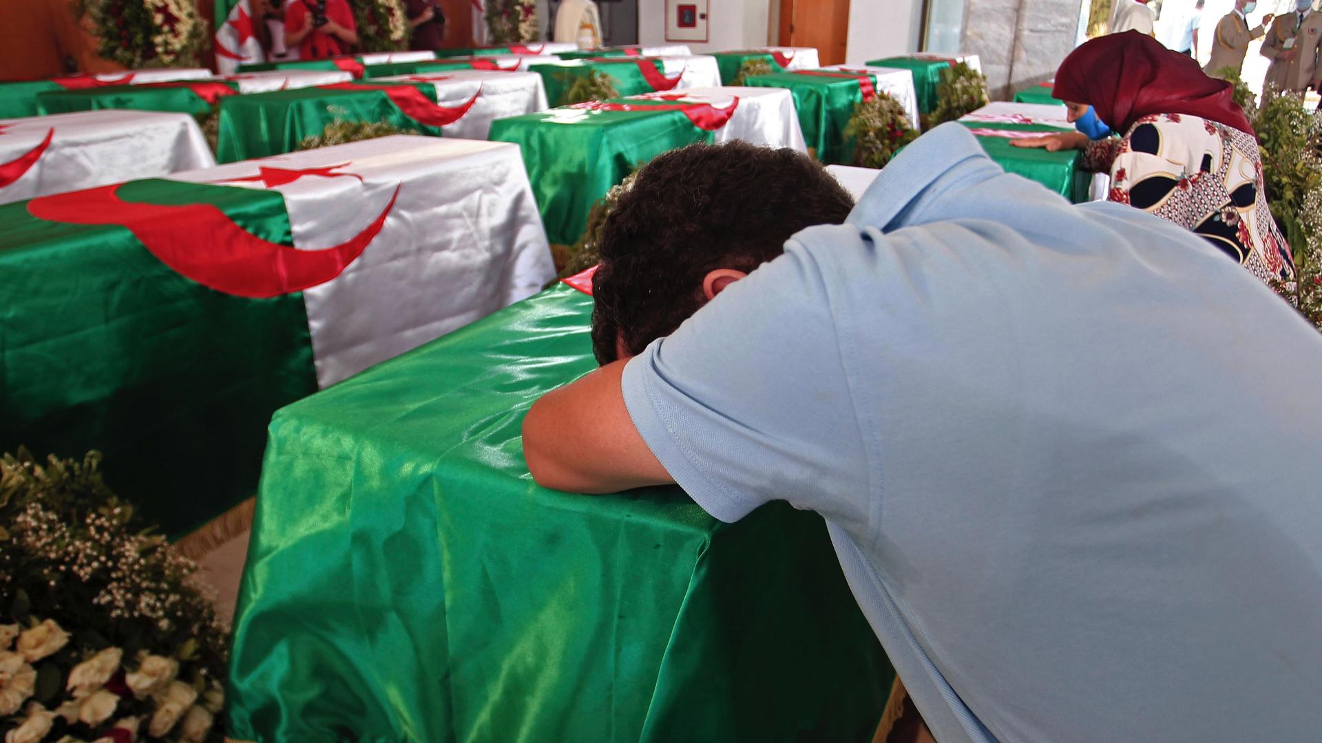 A man wearing a blue shirt leans his body over a coffin draped with the Algerian flag in a room full of coffins. 