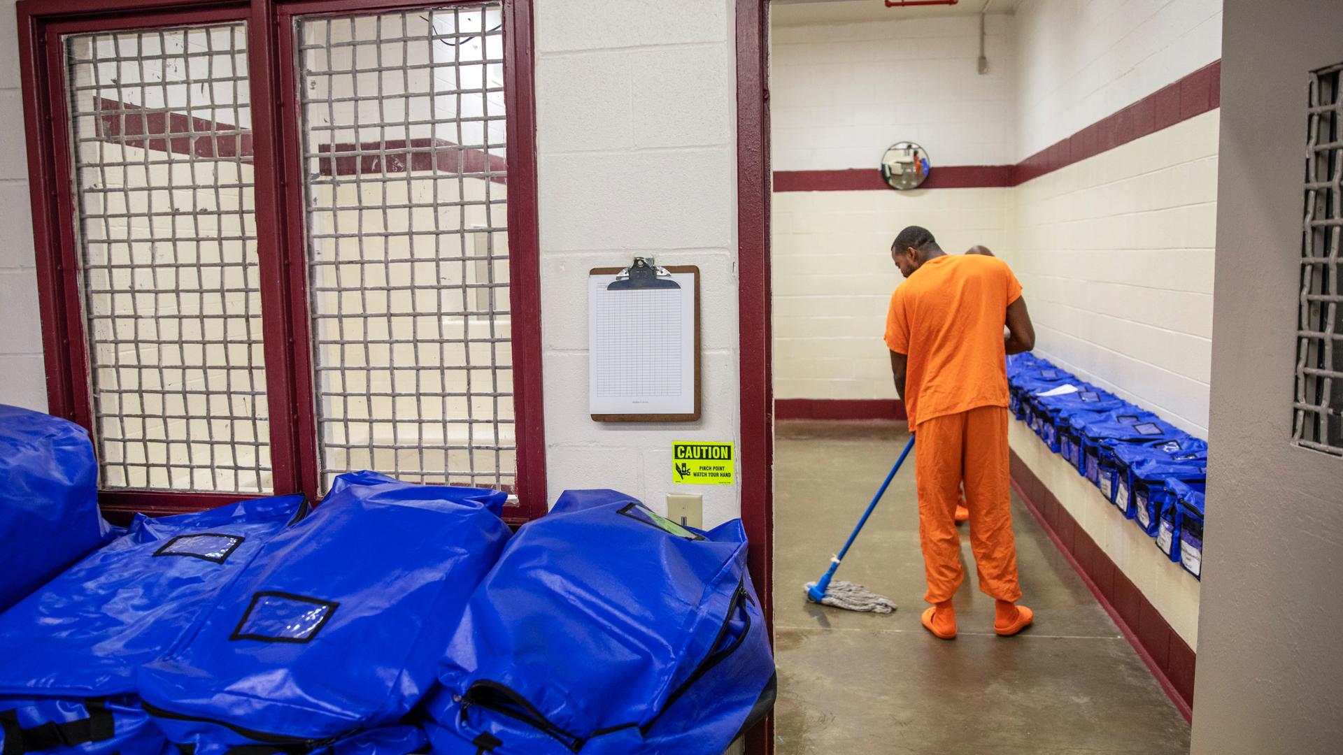 An inmate wearing an orange suit mops near blue plastic bags inside a detention center. 