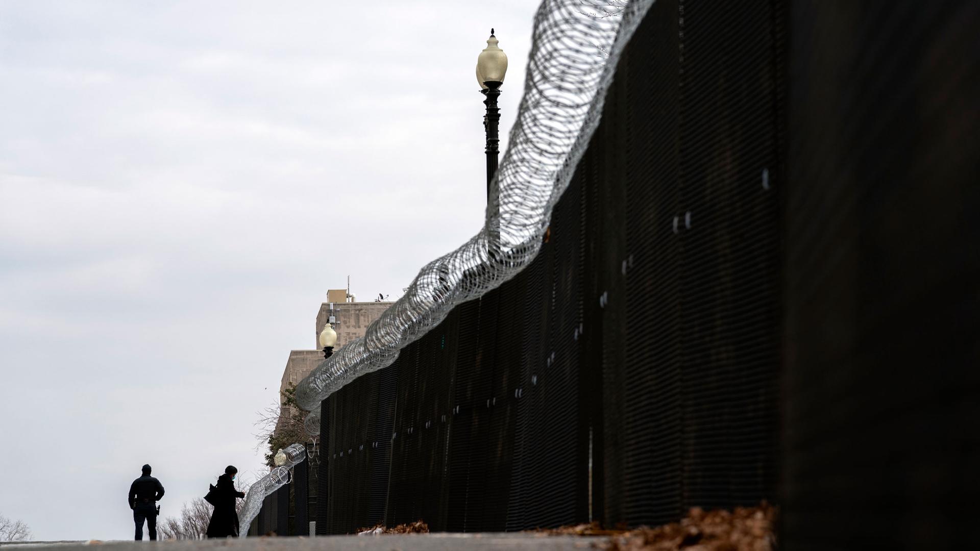 A tall fence is shown with barbed wire running along the top and a dark sheet obstructing the view through with two people shown in the distance.