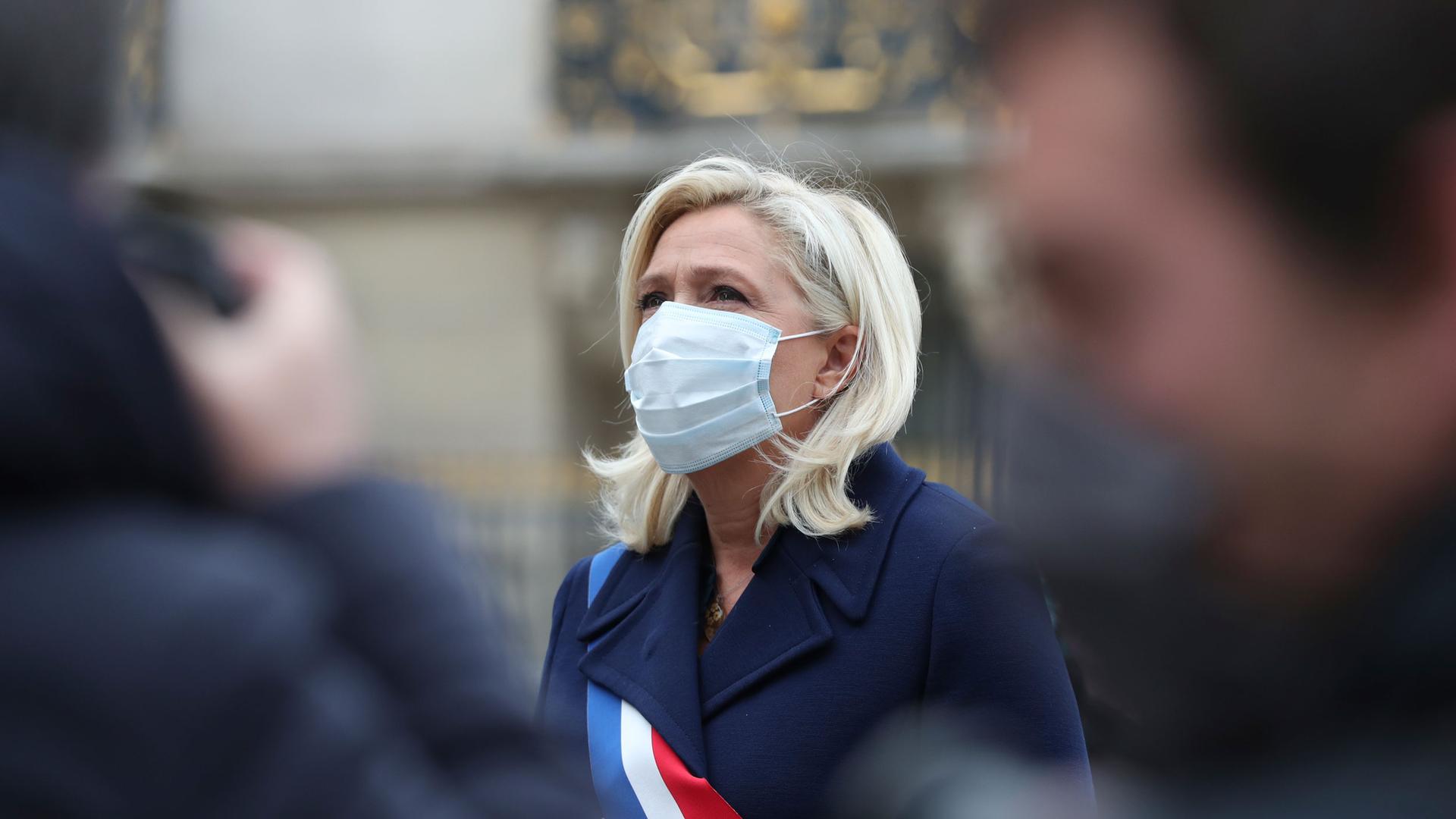 Marine Le Pen is shown wearing a blue jacket and a sash with the colors of the French flag over her shoulder.