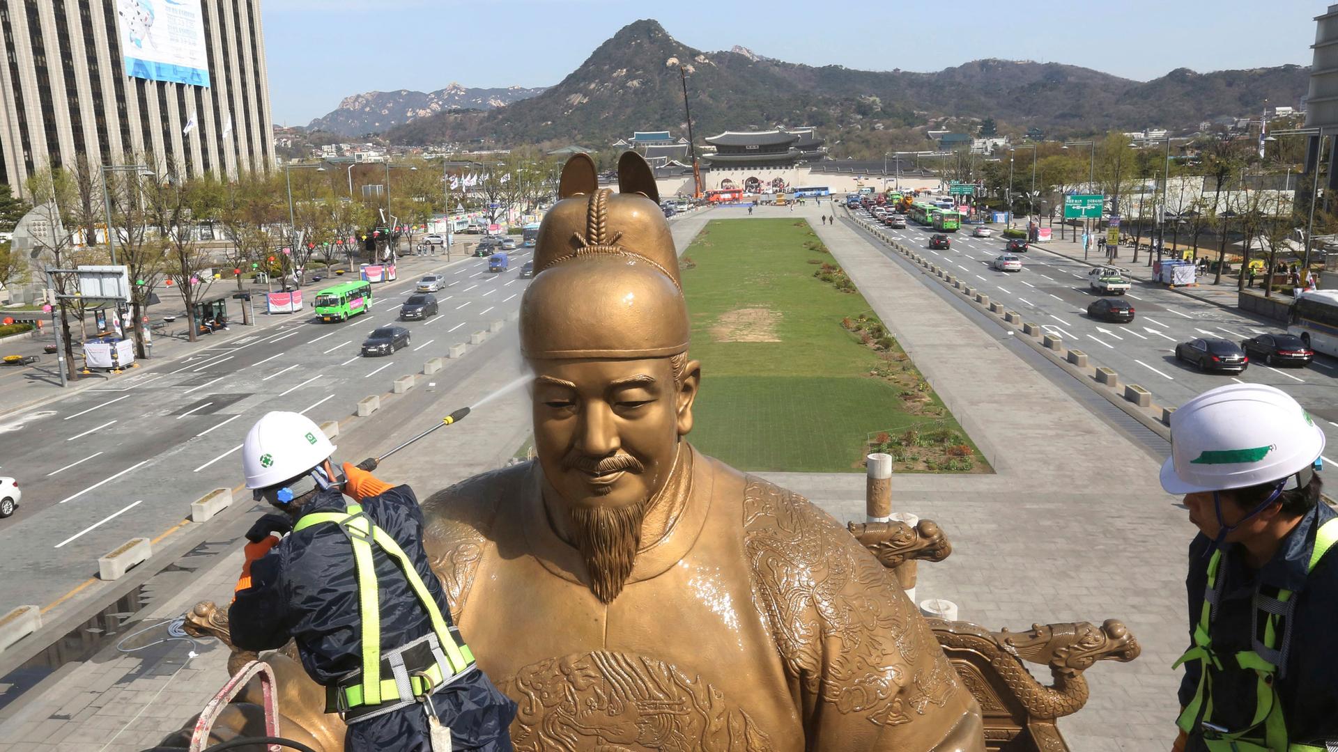 A worker gives a spring cleaning to the large gold-colored statue of King Sejong with mountains shown in the distance.