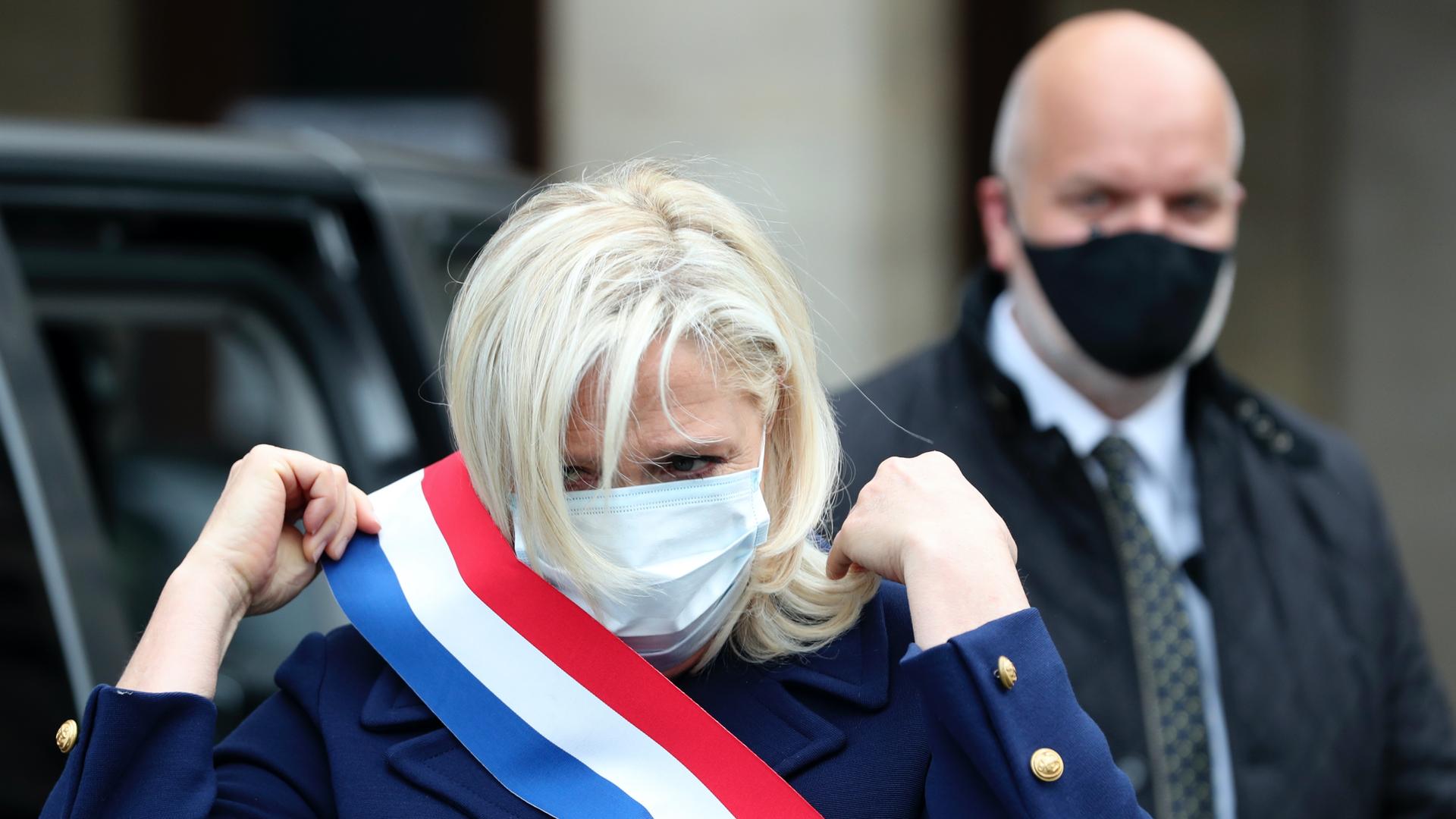 A white woman wearing a face mask also wears a red, white and blue sash around her chest and a man with a black mask stands behind her.