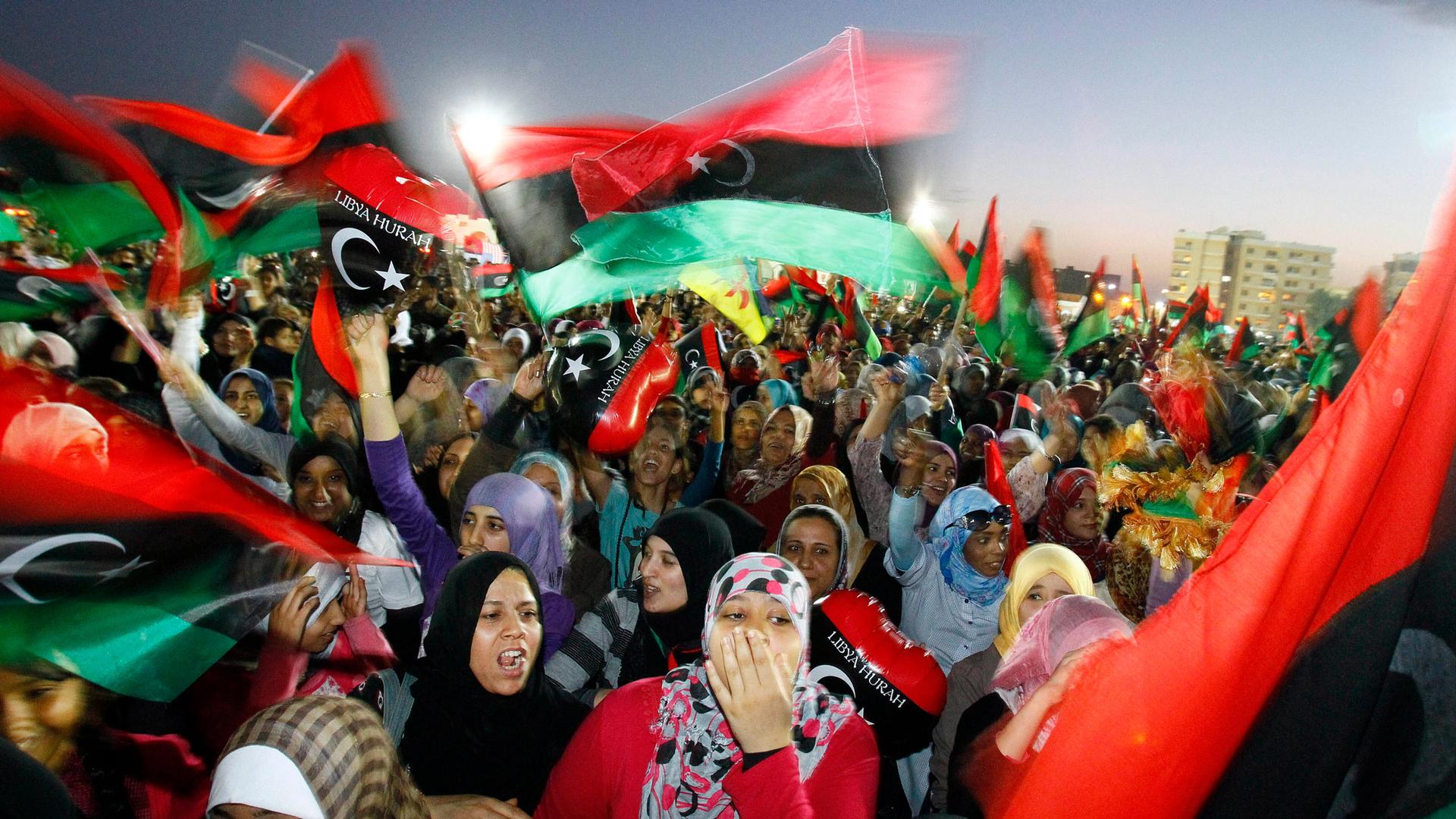 A large crowd of people are shown with many carrying the Libyan flag in a blurred motion photograph.