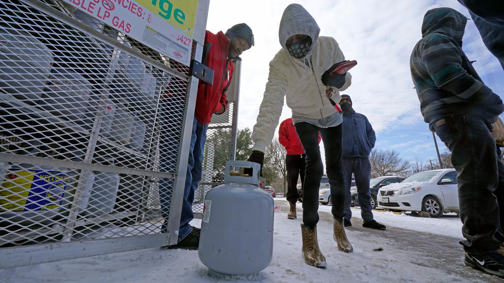 A woman is shown bending over with a white propane tank in her hand and wearing a hooded jacket.
