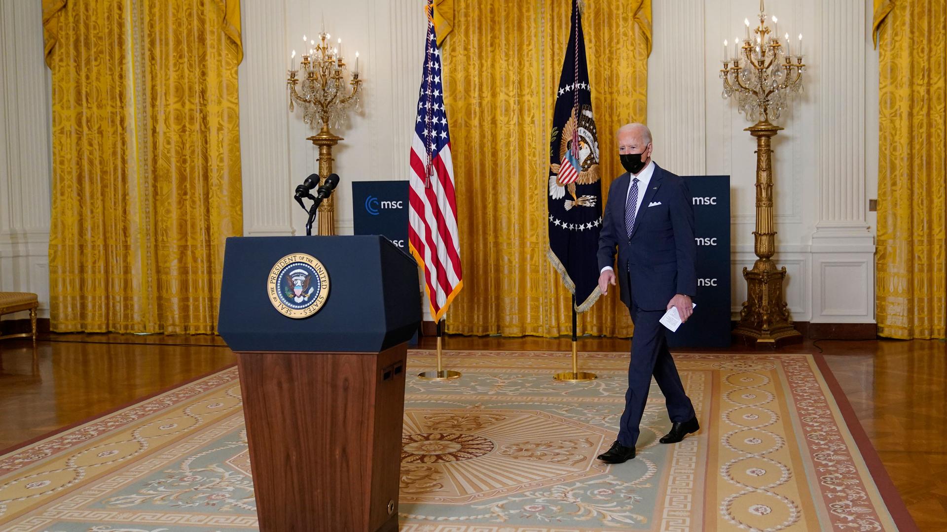 President Joe Biden is shown walking toward a podium with the White House crest on it and carrying a white piece of paper.