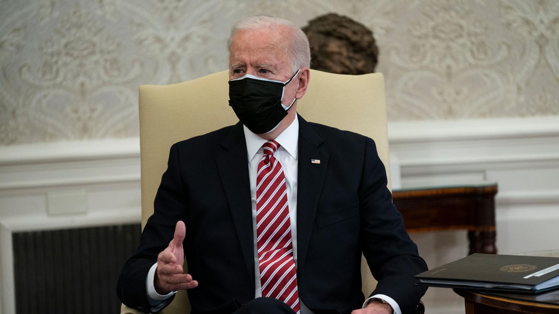 President Joe Biden wearing a dark suit and red and white tie along with a face mask while sitting.