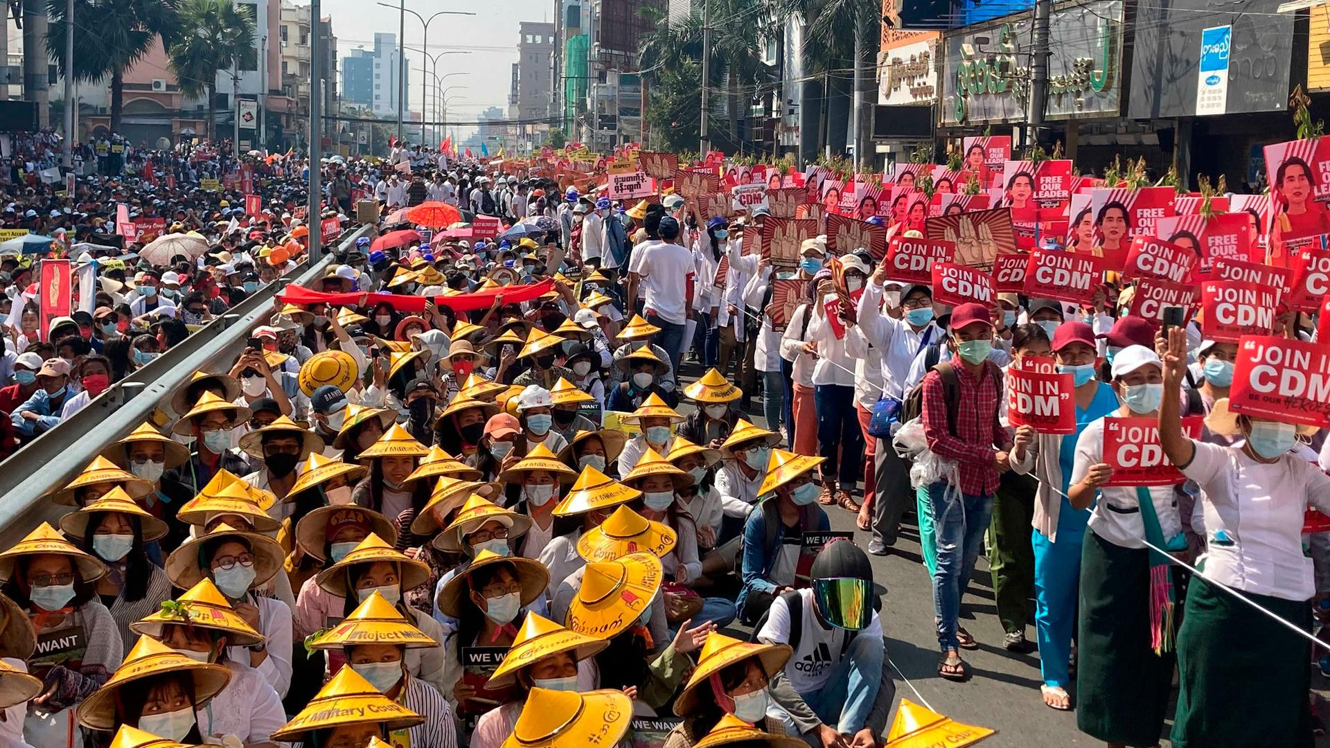 A large crowd of people are shown filling a street with many wearing traditional Asian hats and carrying red signs.