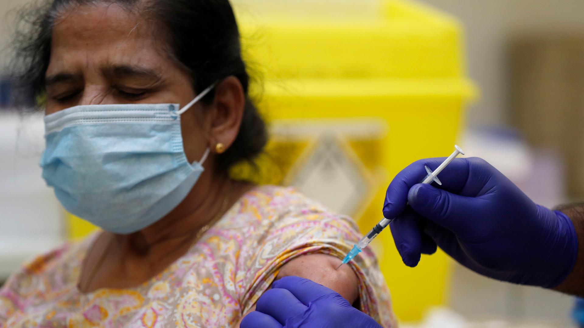 An Indian British woman wears a face mask and receives a shot in her arm with someone wearing purple gloves. 