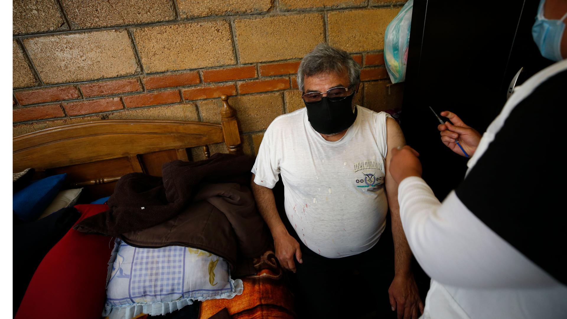 A nurse administers a shot to an elderly man wearing a white T-shirt and black pants at his home. 