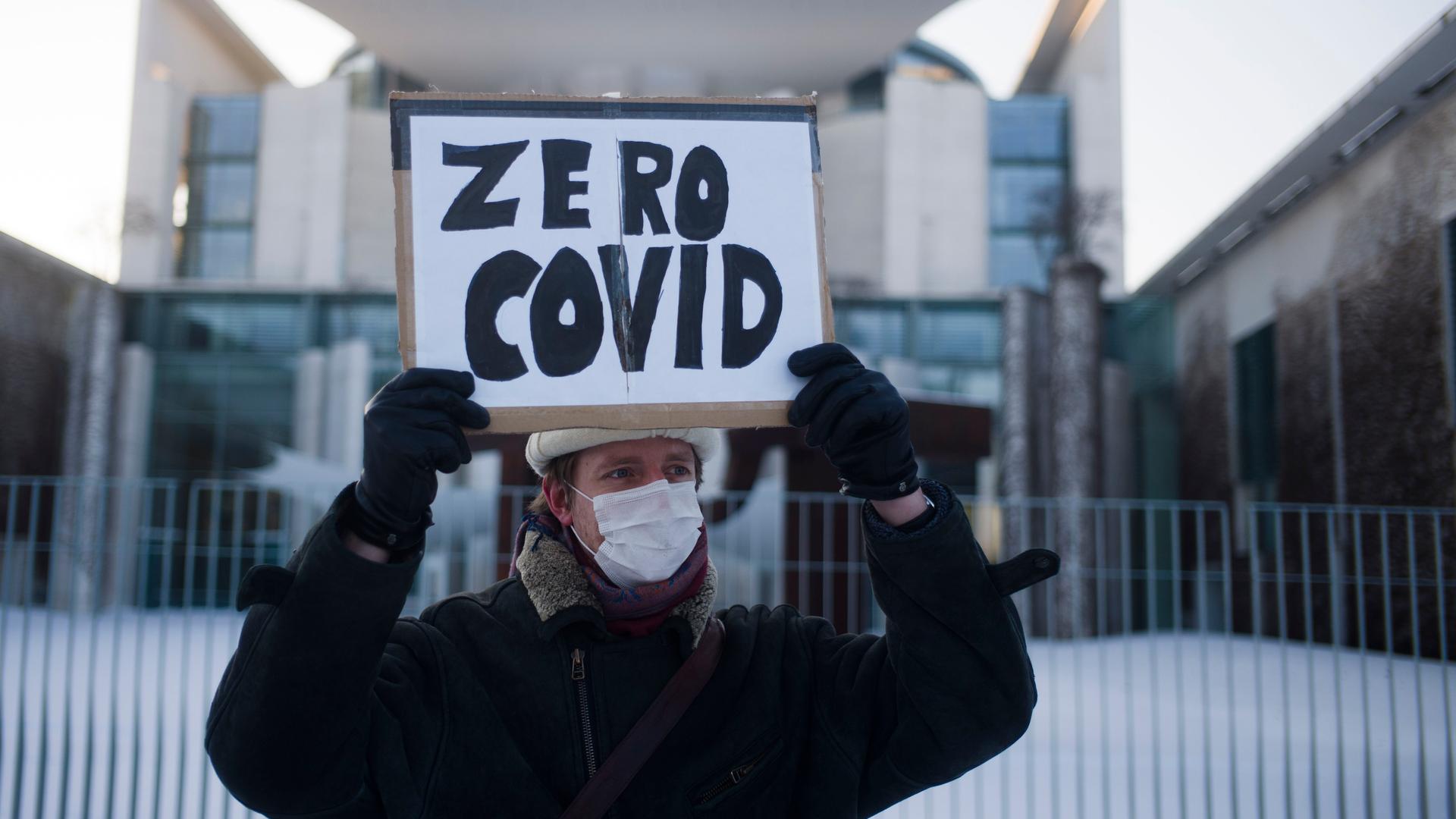 A man holds a sign that says "zero COVID" wearing a winter jacket and hat outside in front of chancellery in Berlin. 
