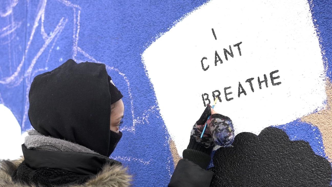A person paints a sign that says "I can't breathe" on a wall with a purple background. 