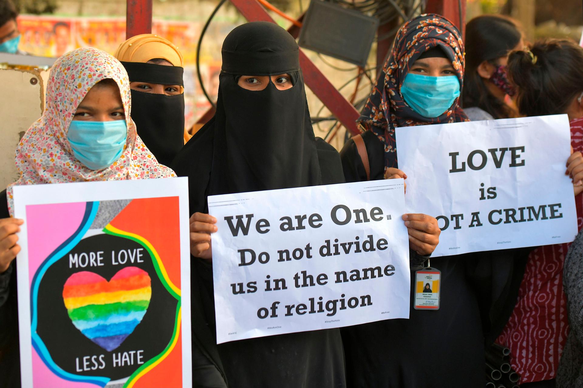 Three women protestors hold up signs reading "More Love, Less Hate"; "We are One. Do not divide us in the name of religion"; and "Love is not a crime."