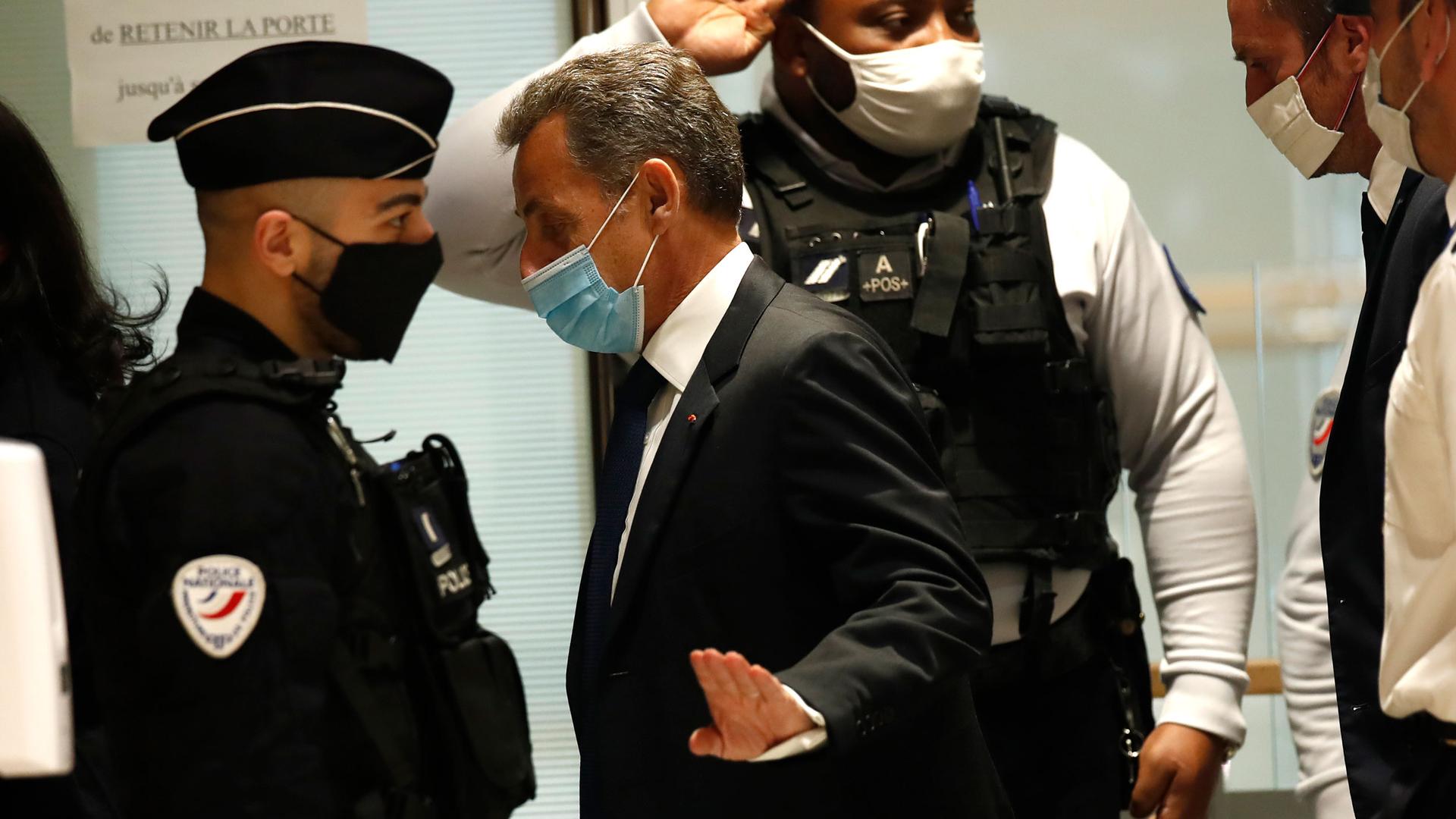 Former French President Nicolas Sarkozy is shown wearing a dark suit and tie with a face mask, walking past security officials.