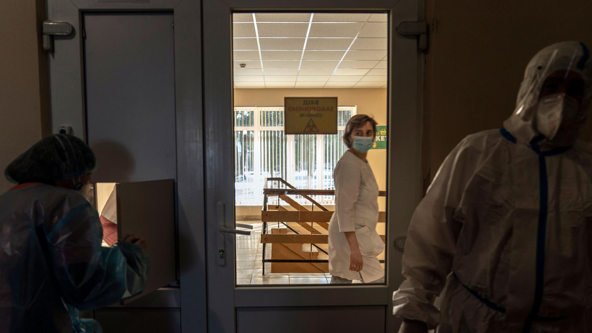 A nurse is shown through the glass of a closed door looking into a darkened room with two other medical professionals dressed in protective clothing.