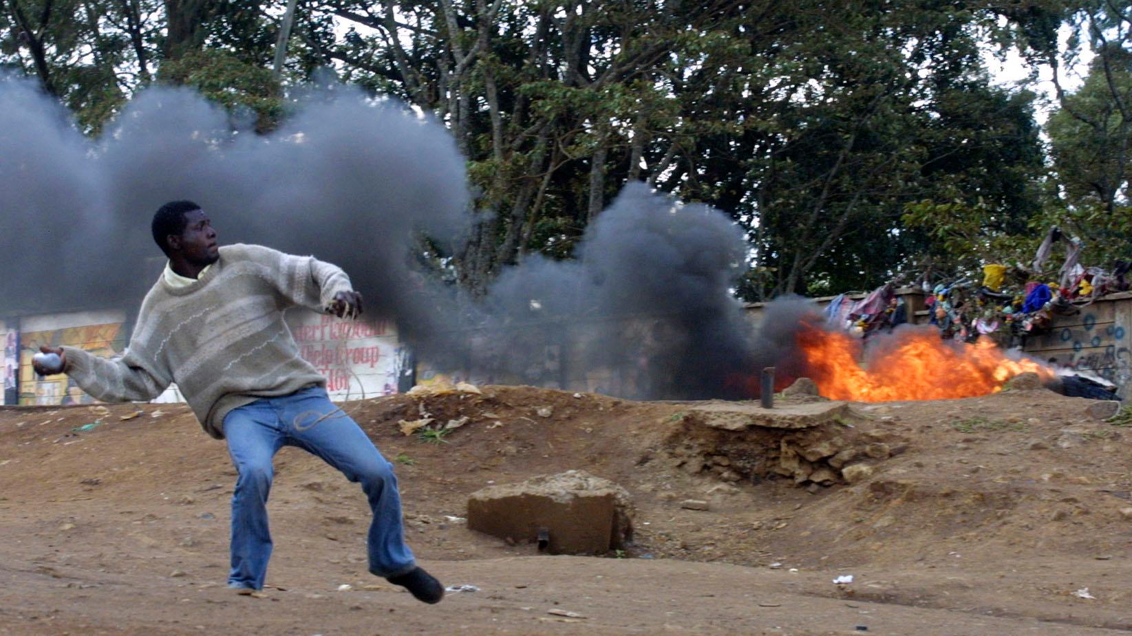 A man wearing jeans and sweater throws a rock near a fire burning outside