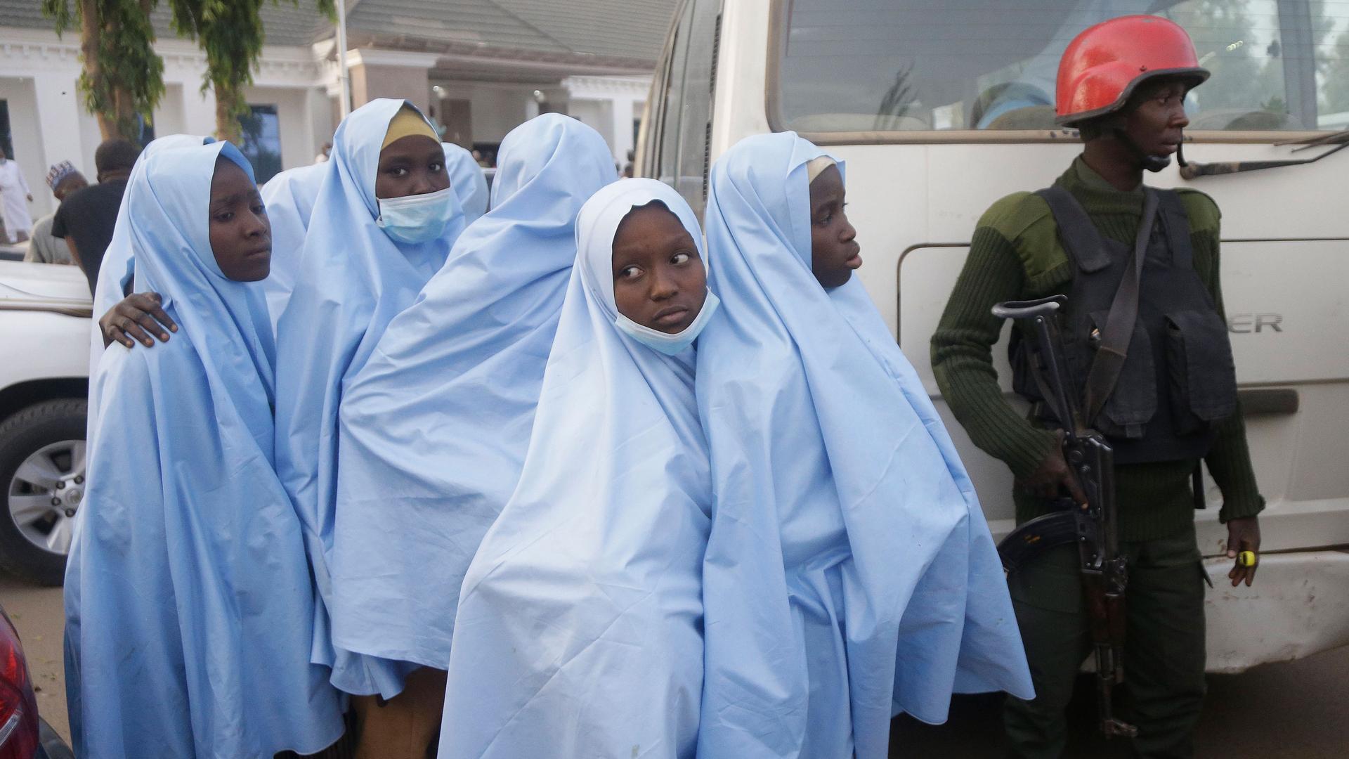 Several young school children are shown wearing blue traditional Muslim coverning with a security official hearing a red helmet standing by. 