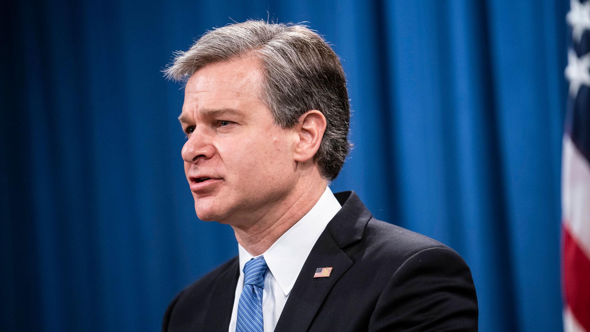 FBI Director Christopher Wray is shown wearing a dark suit and blue striped tie with a US flag pin on his lapel.