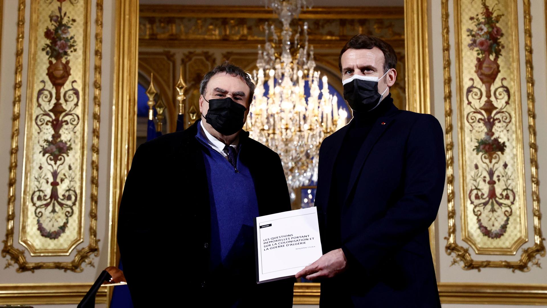French President Emmanuel Macron is shown standing next to French historian Benjamin Stora, both men wearing dark jackets and Macron holding a white paper.