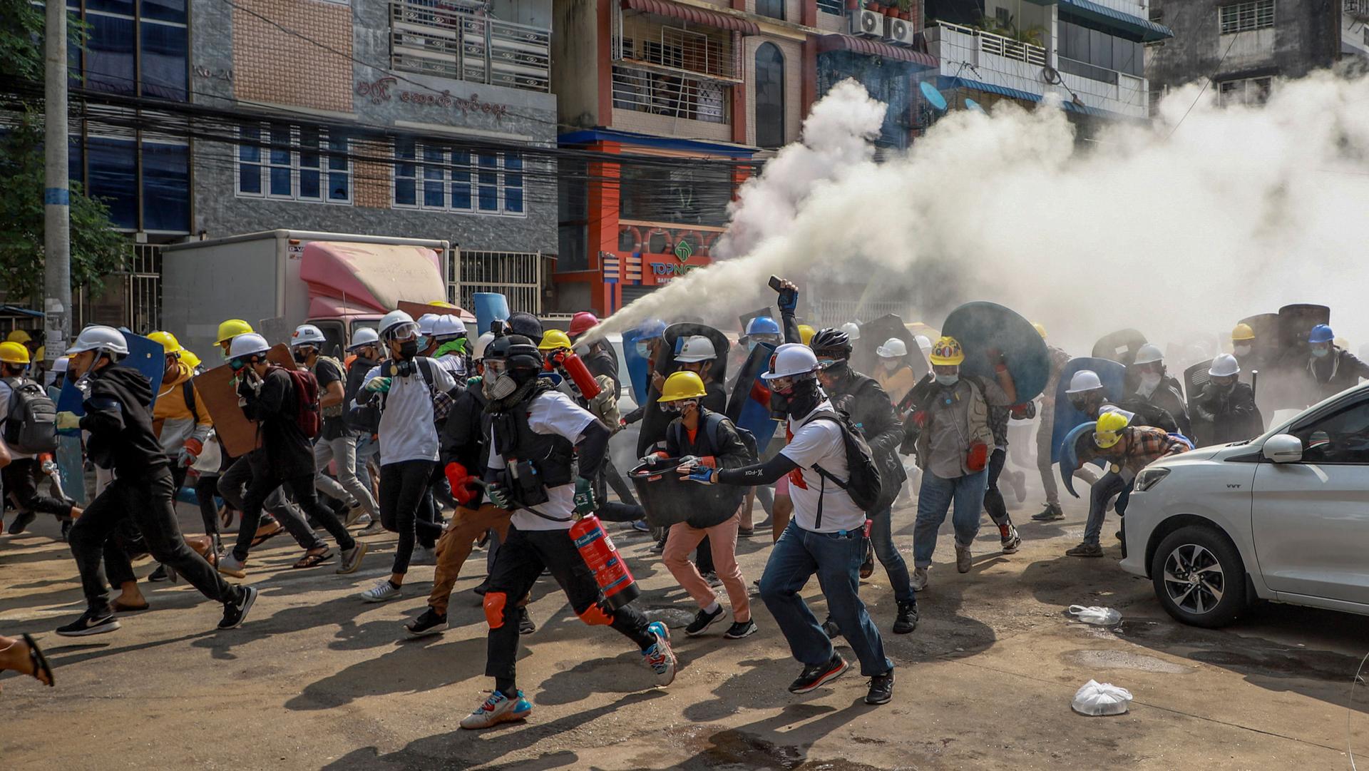 A large crowd of people are shown, many wearing construction hard hats, as one of them sprays a fire extinguisher into the air in the street.