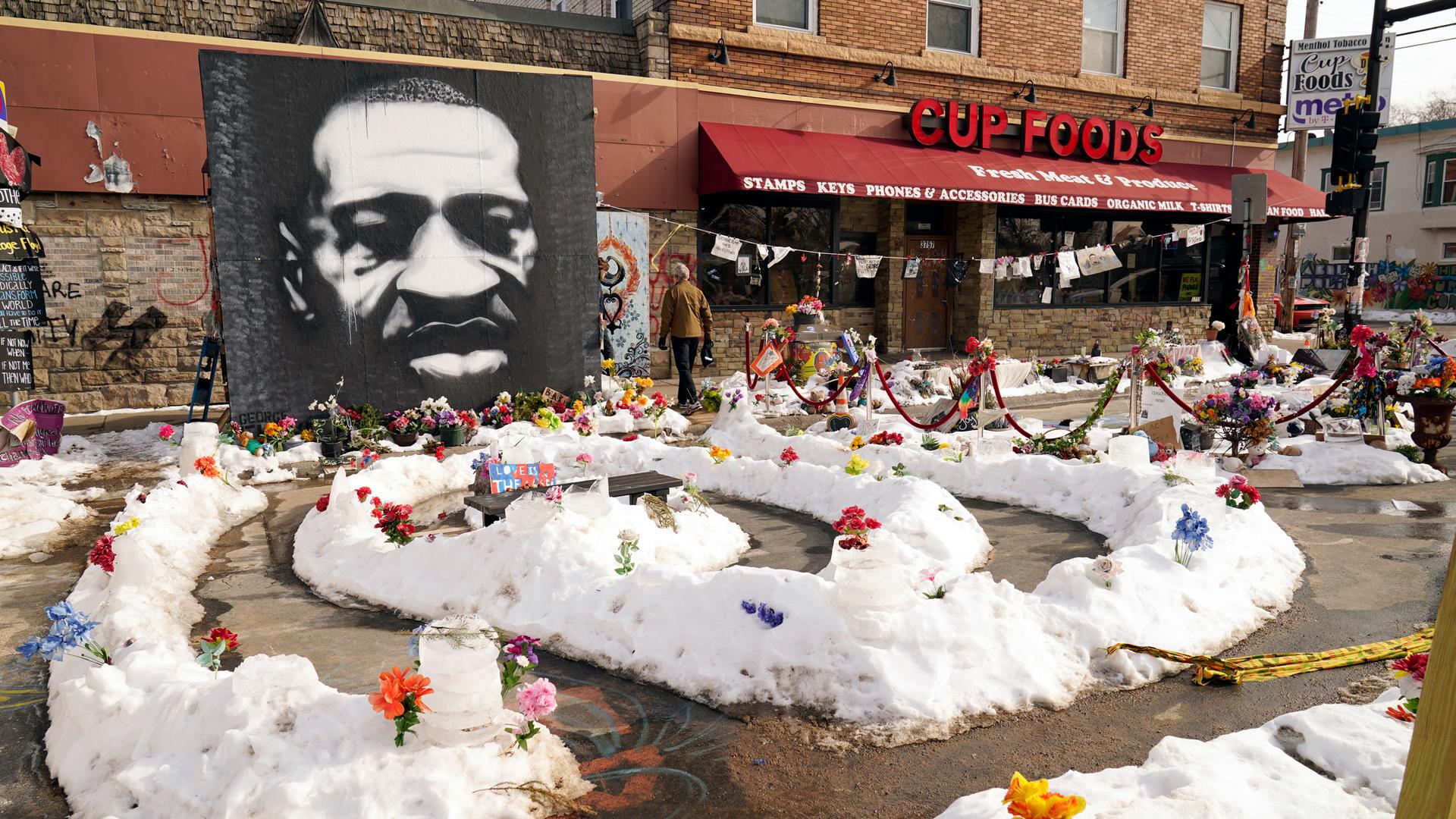 A large black and white portrait of George Floyd is shown afixed to the wall of a Cup Foods store with flowers placed in snow piles.