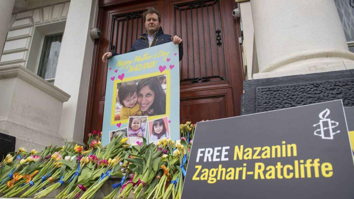 Richard Ratcliffe is shown standing in front of large wooden doors and holding a large card with a picture of Nazanin Zaghari-Ratcliffe on with with dozens of flowers nearby.
