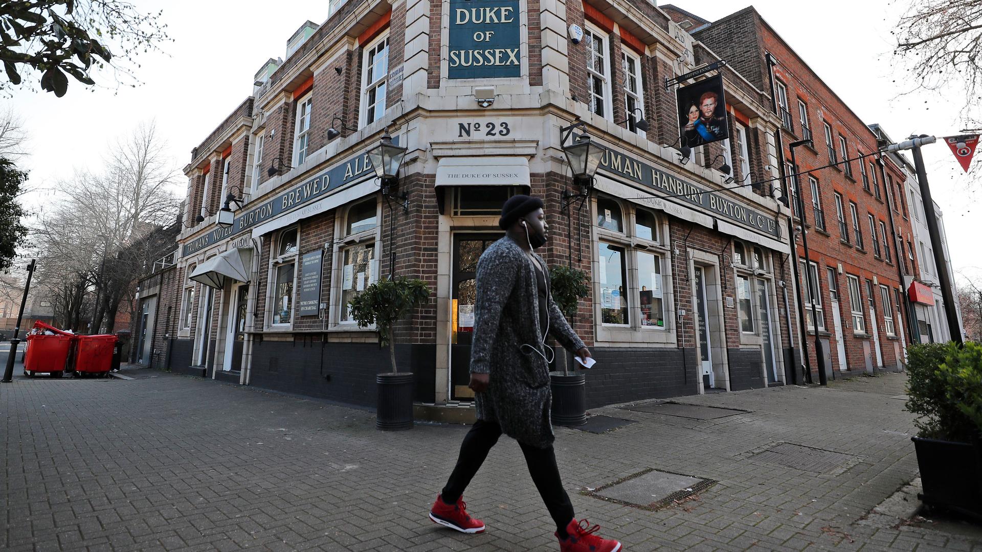 A man is shown with white headphones and read shoes walking past a pub called The Duke of Susex.