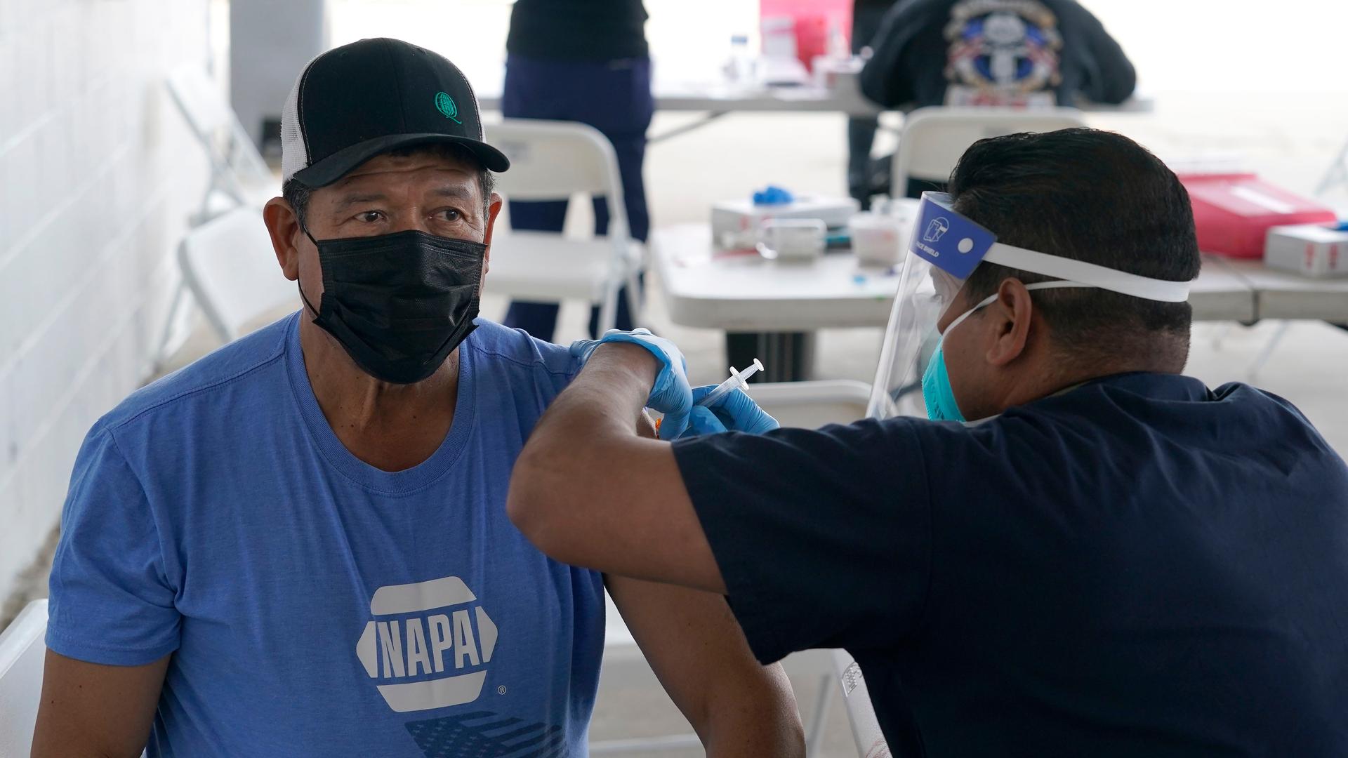 A man wearing a blue shirt and black cap gets a vaccine shot by a health care worker. 