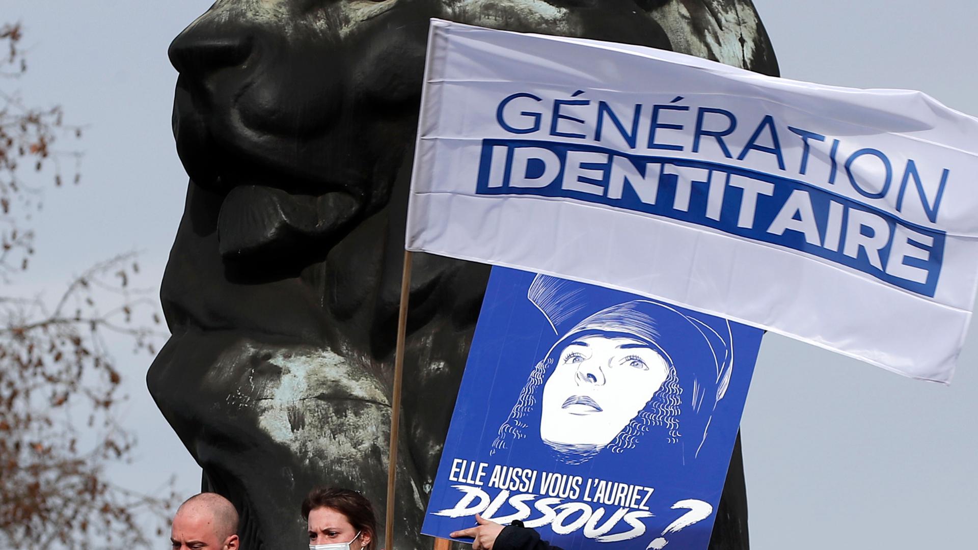 Two masked people hold a blue and white sign with "Generation Identity" written on it. 
