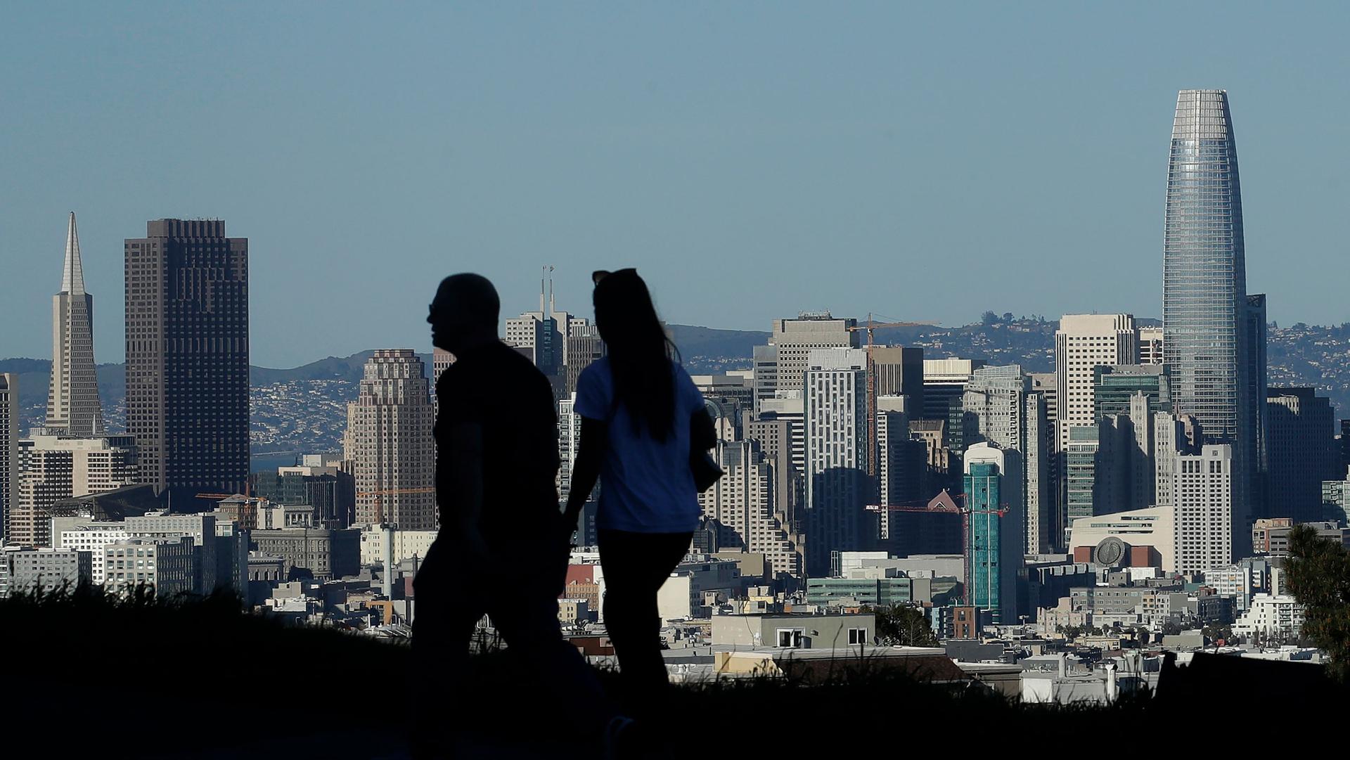 The downtown skyline of San Francisco is shown in the distance with a couple walking in the nearground in shadown.