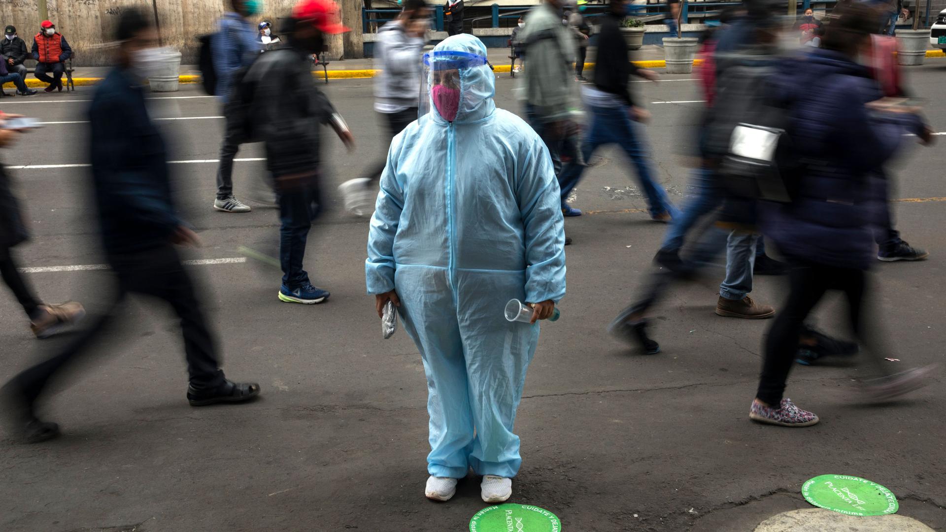 A woman is shown standing and wearing a full blue medical protective outfit with several people walking past her in blurred motion.