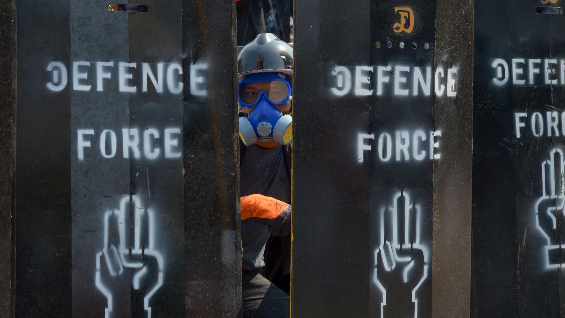 A person wearing an air-filtration mask and helmet is show looking through makeshift metal barriers with the words 'Defense Force' and a hand holding up three fingers painted on them.