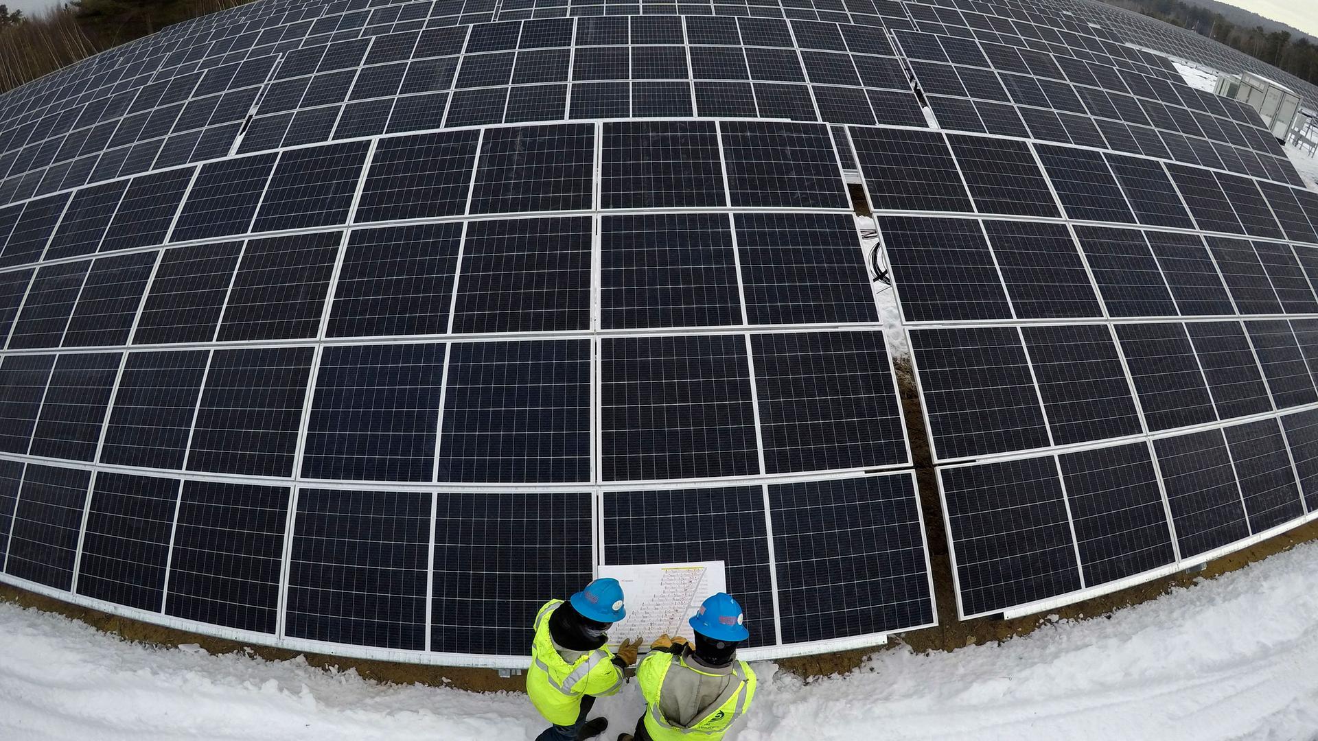 Several rows of large solar panels are shown with a wide angle lens with two people at the bottom of the photo wearing yellow safety vests and hard hats.