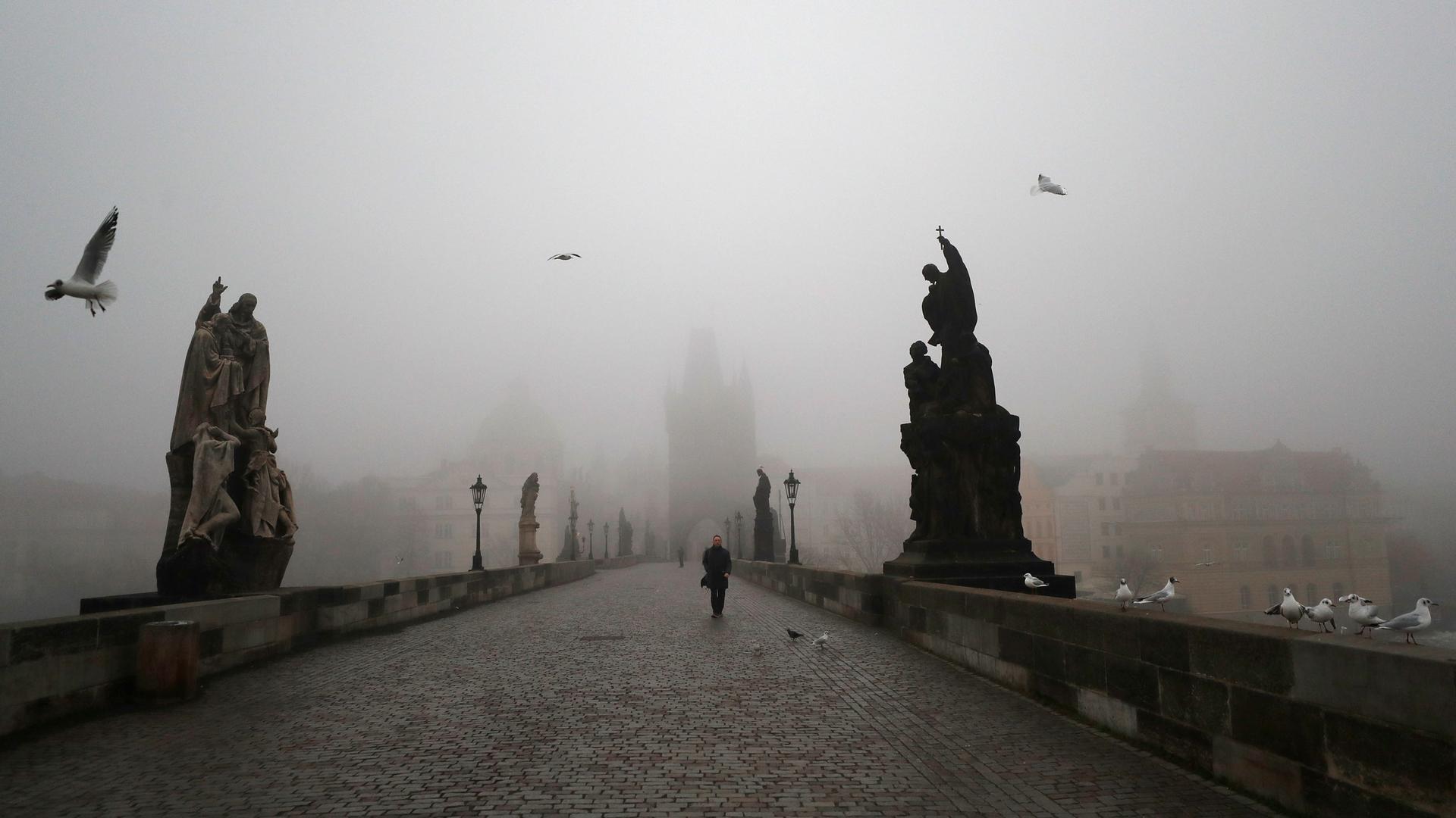A cloudy, gray day is shown from one end of the Charles Bridge, with several stone statues on either side and a man walking in the distance.