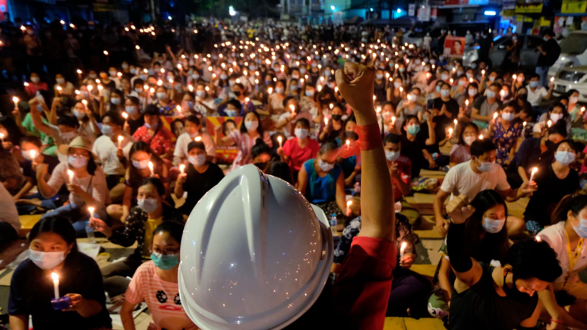 A person is shown in the near ground with their fist in the air and wearing a white construction helmet with a large crowd of people sitting and holding candles.