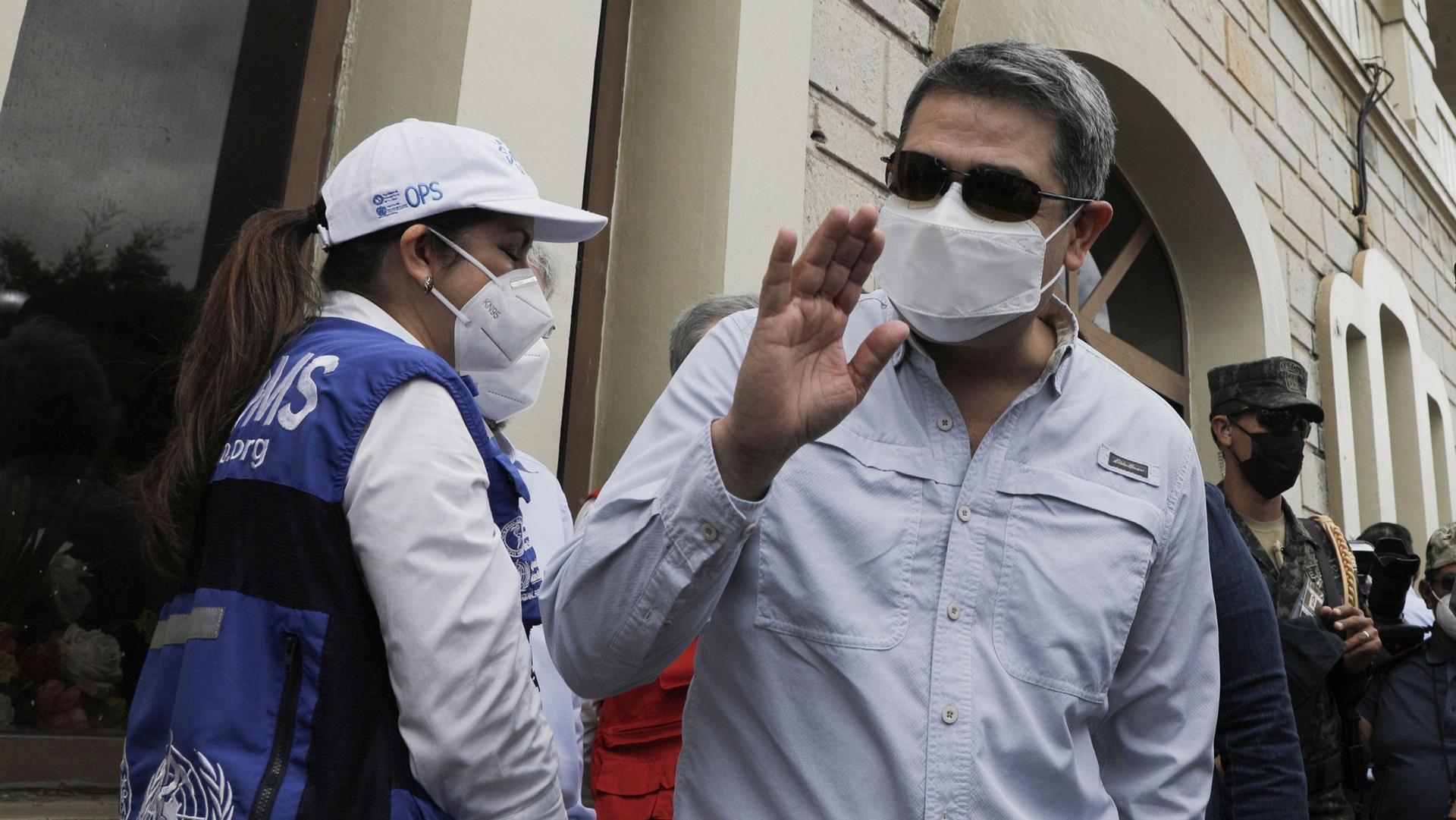 Honduran President Juan Orlando Hernandez is shown wearing a medical mask and sunglasses while waving his right hand.