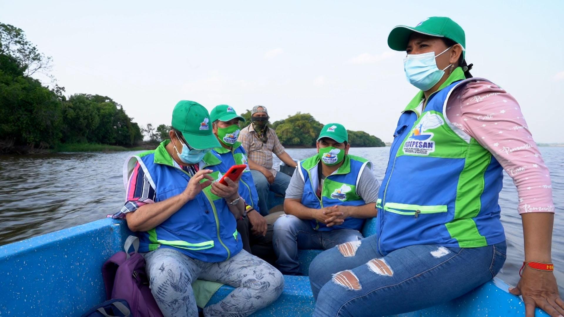 Four people wearing blue jackets and green caps ride in a boat on a lake. 