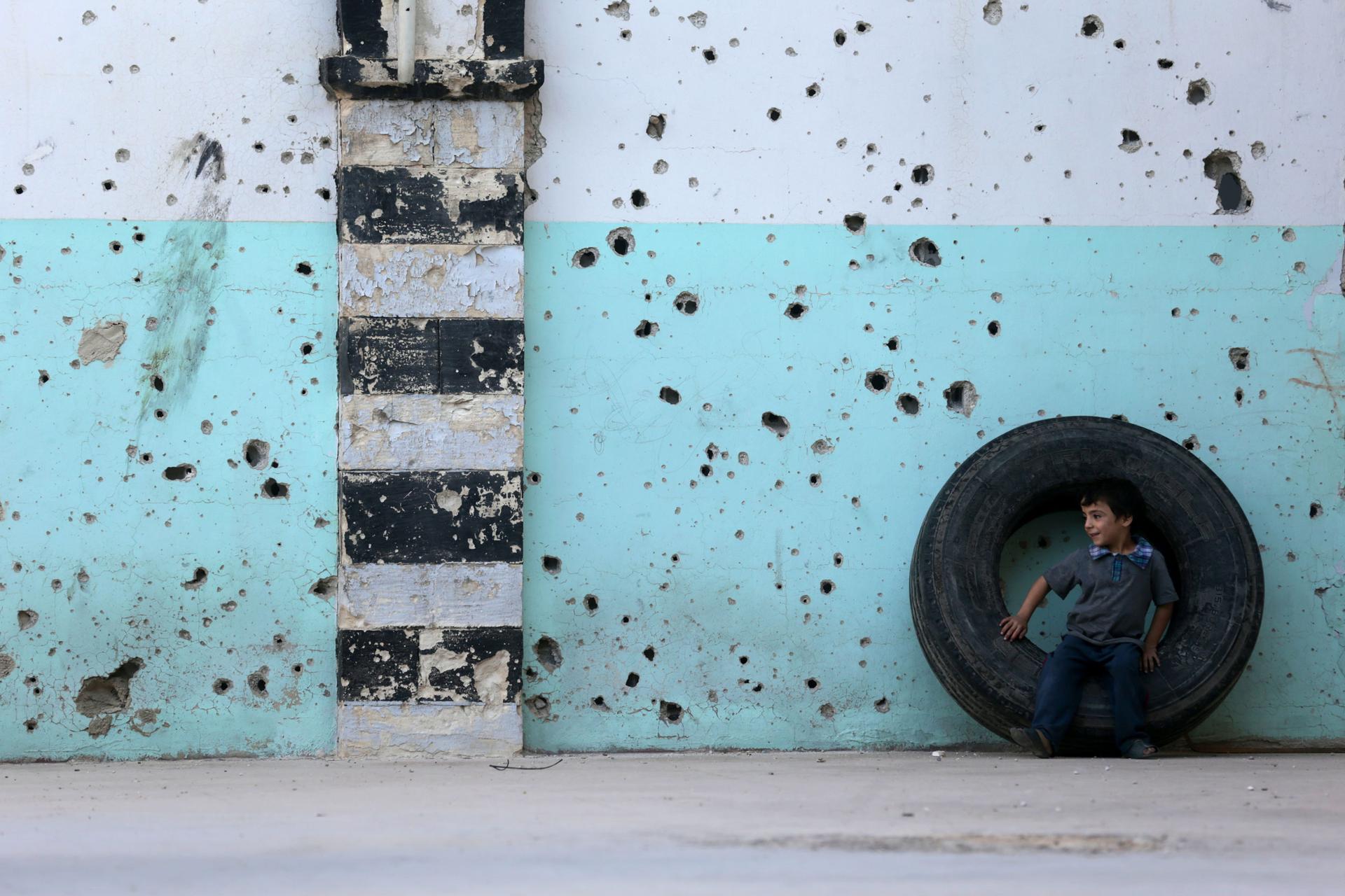A young boy is shown sitting in a large tire against a blue wall riddled with bullet holes.