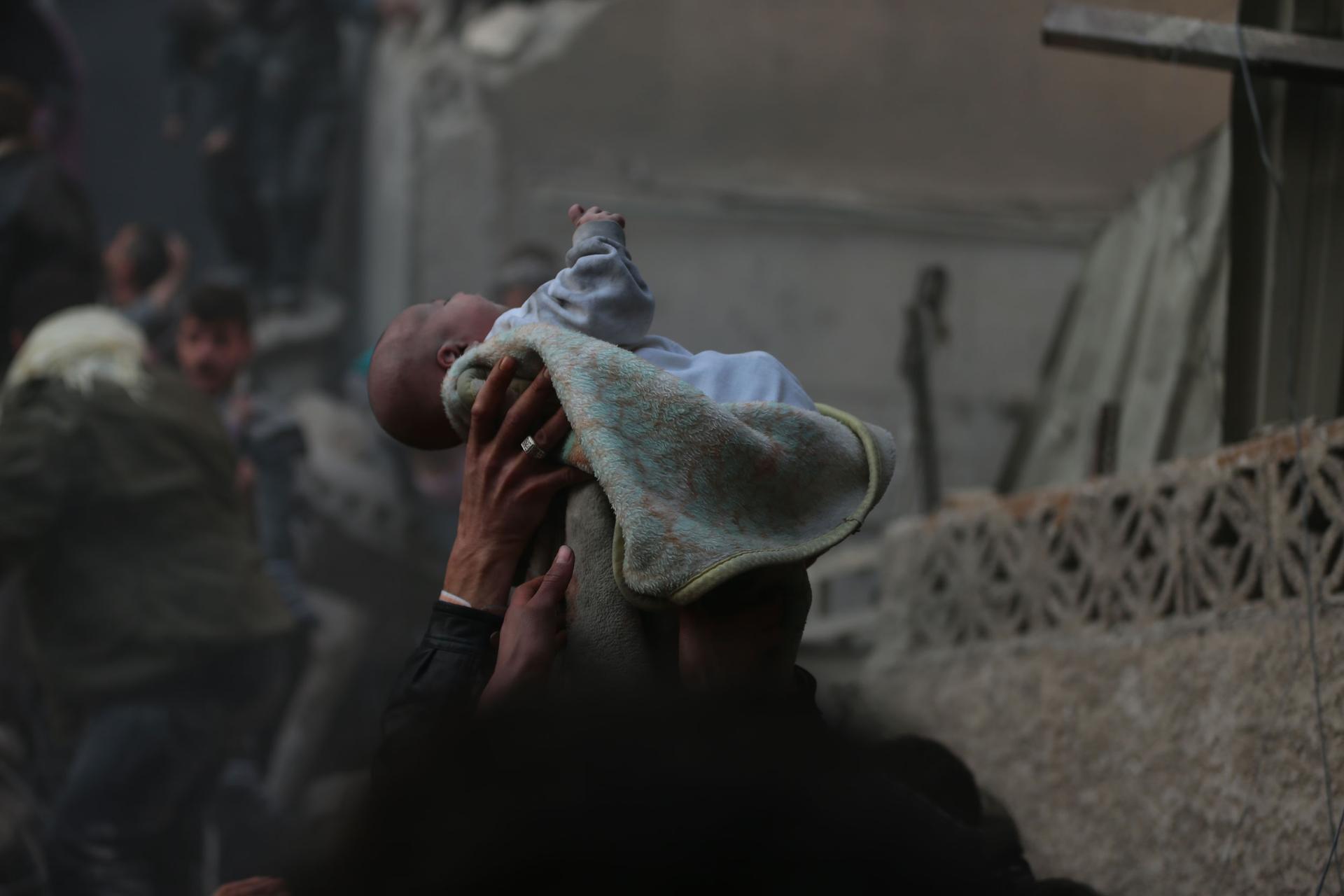 A small baby is shown wrapped in blanked and held above the heads people amid blackened walls of buildings.