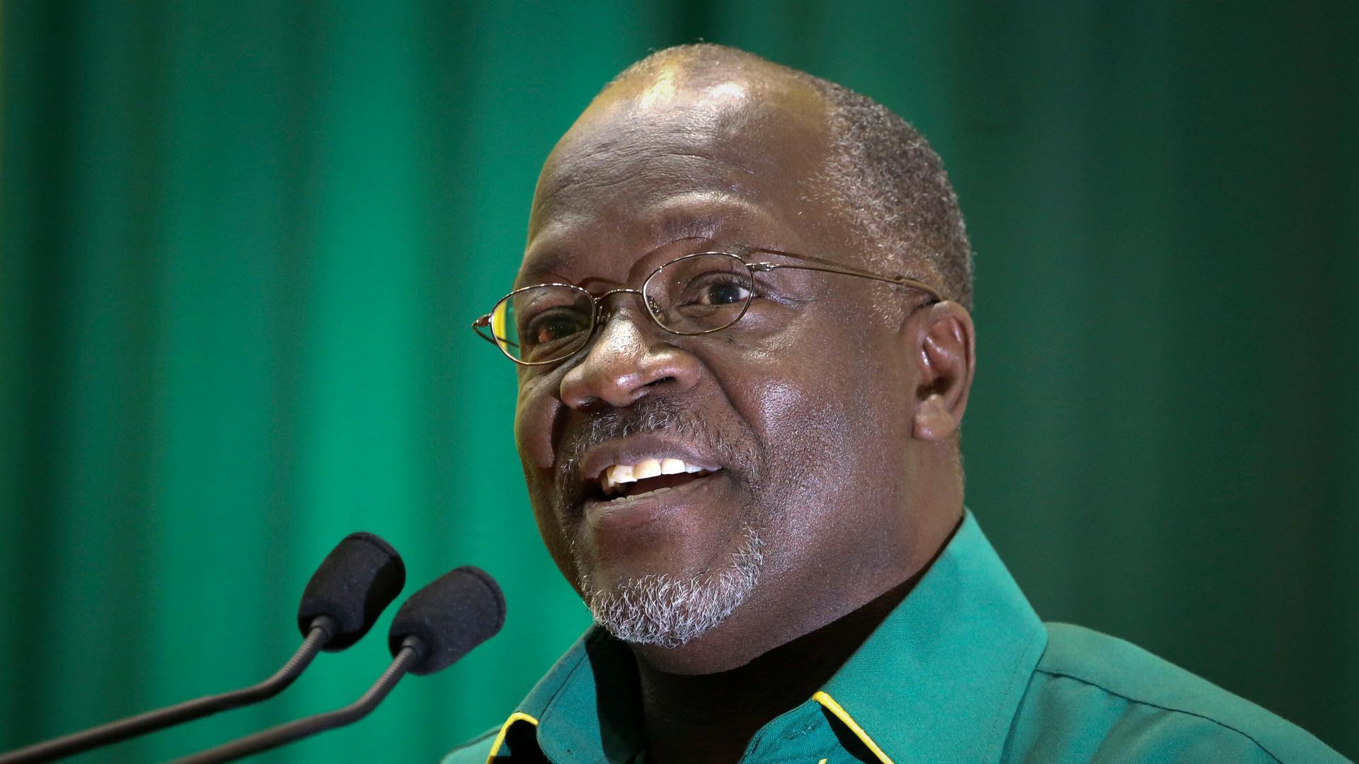 John Magufuli wears glasses a green shirt and speaks at a podium with a green background 
