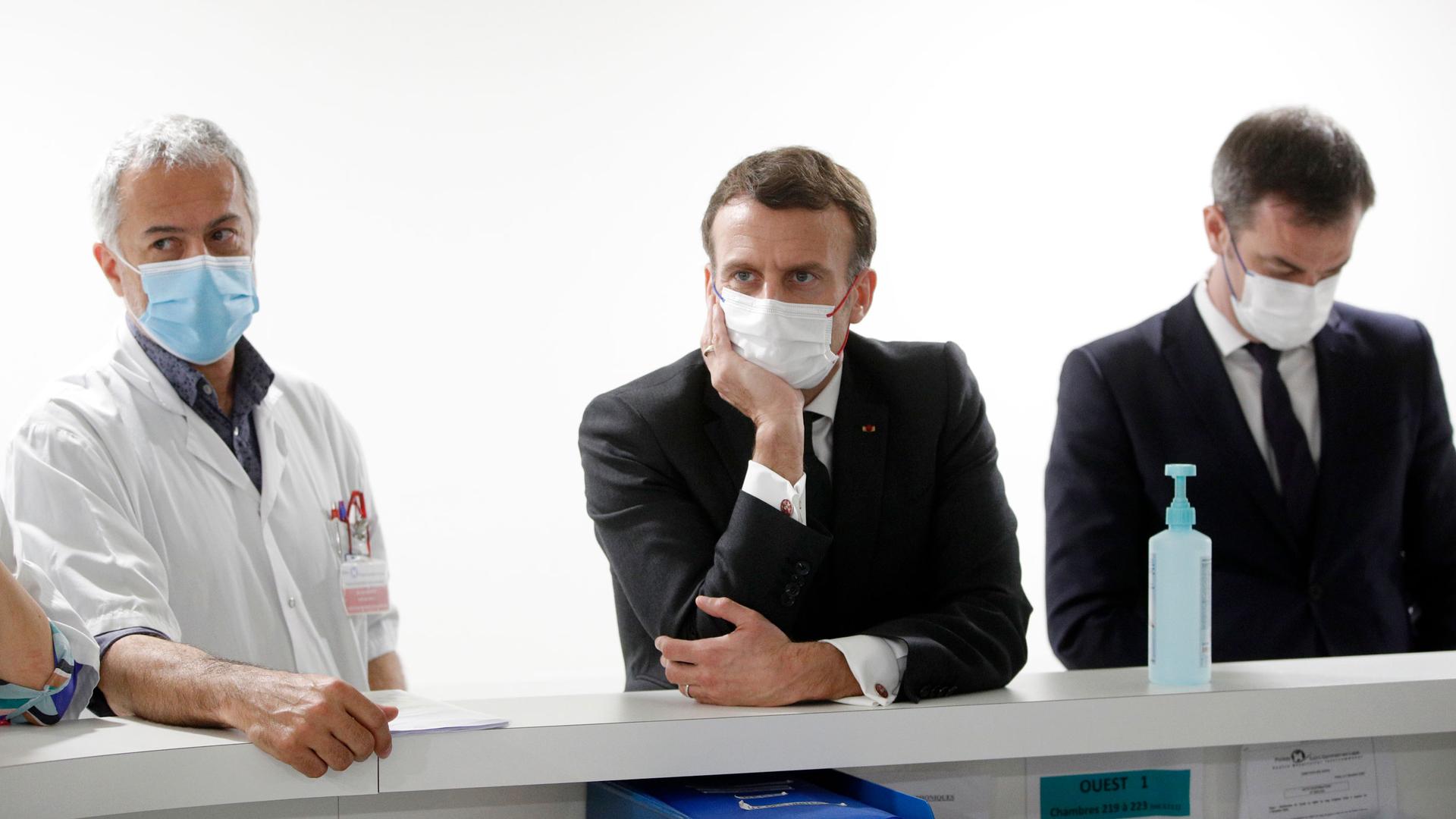 French President Emmanuel Macron is shown wearing a dark suit and white face mask while flanked by other officials.