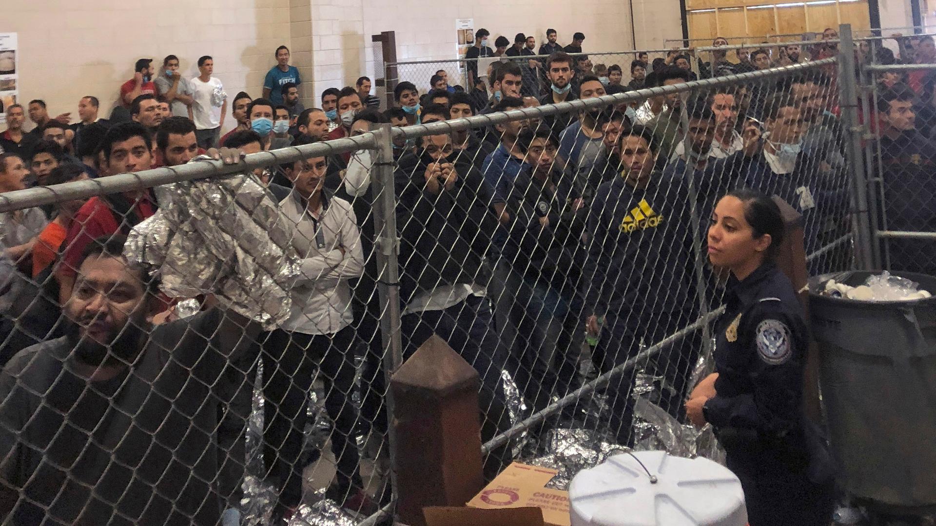 A group of men stand behind a fenced area within a detention center at the border.