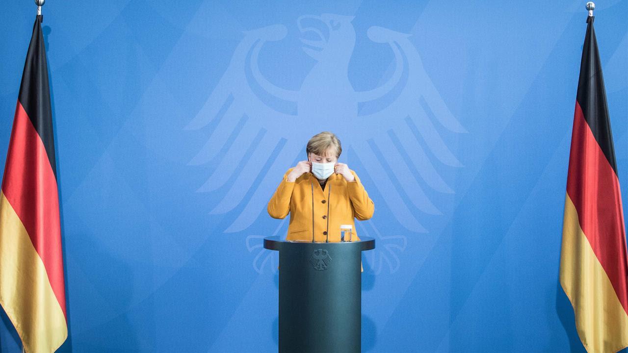 Merkel stands at a podium flanked by two German flags. 