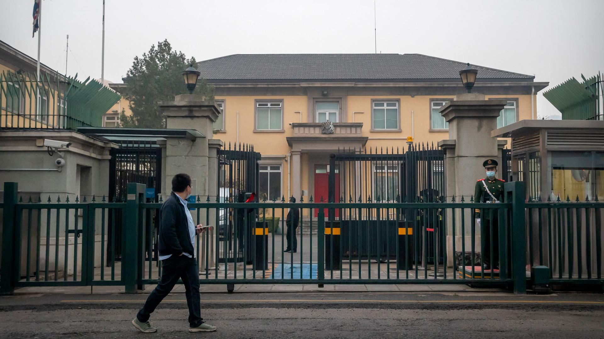 The British Embassy is shown in the distance through a closed metal gate with a Chinese paramilitary police officer standing guard.