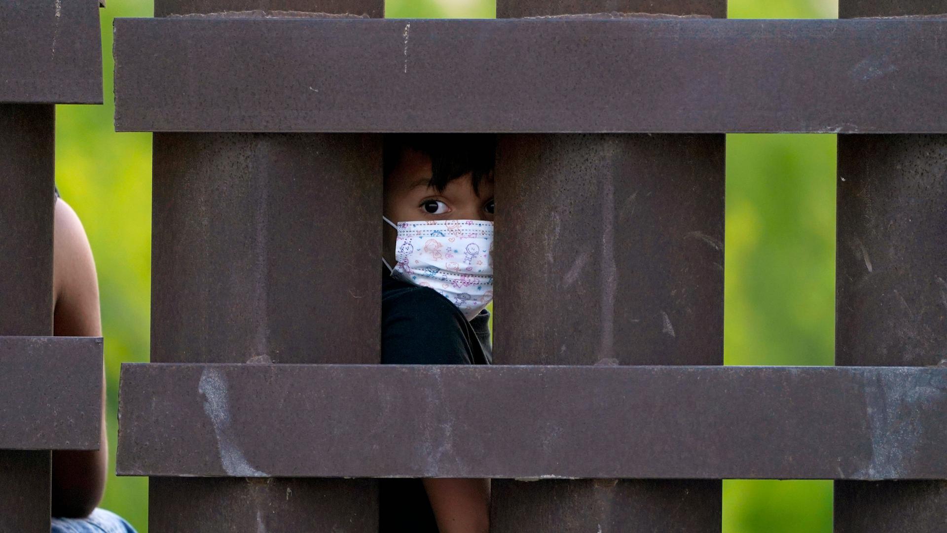 A small child is shown wearing a protective face mask and looking through the large medal polls of the US-Mexico border fence.