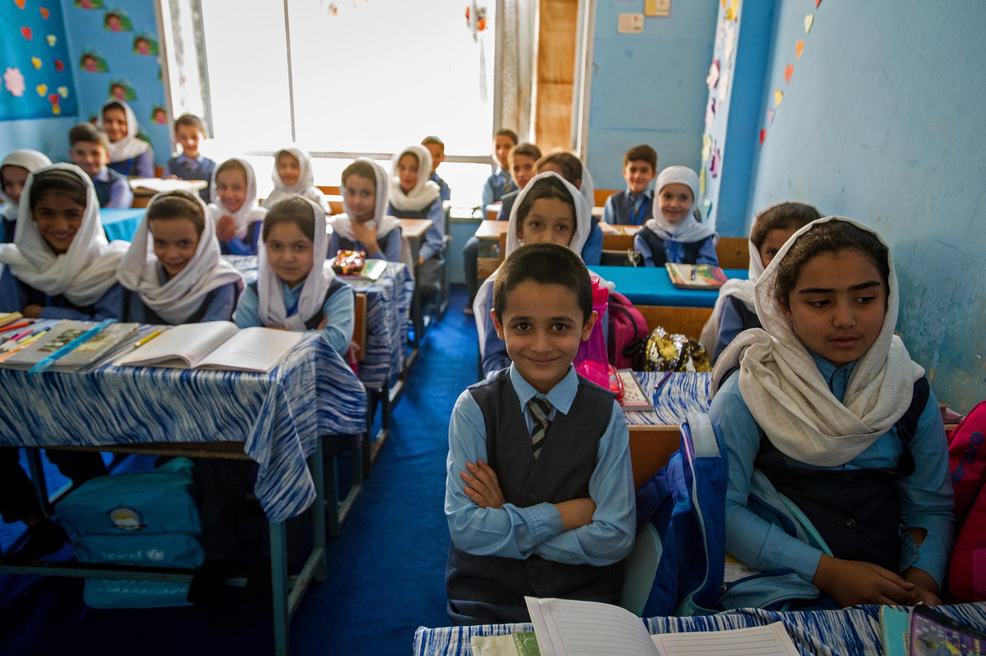 A group of children in a classroom smile for the camera while sitting at their desks.