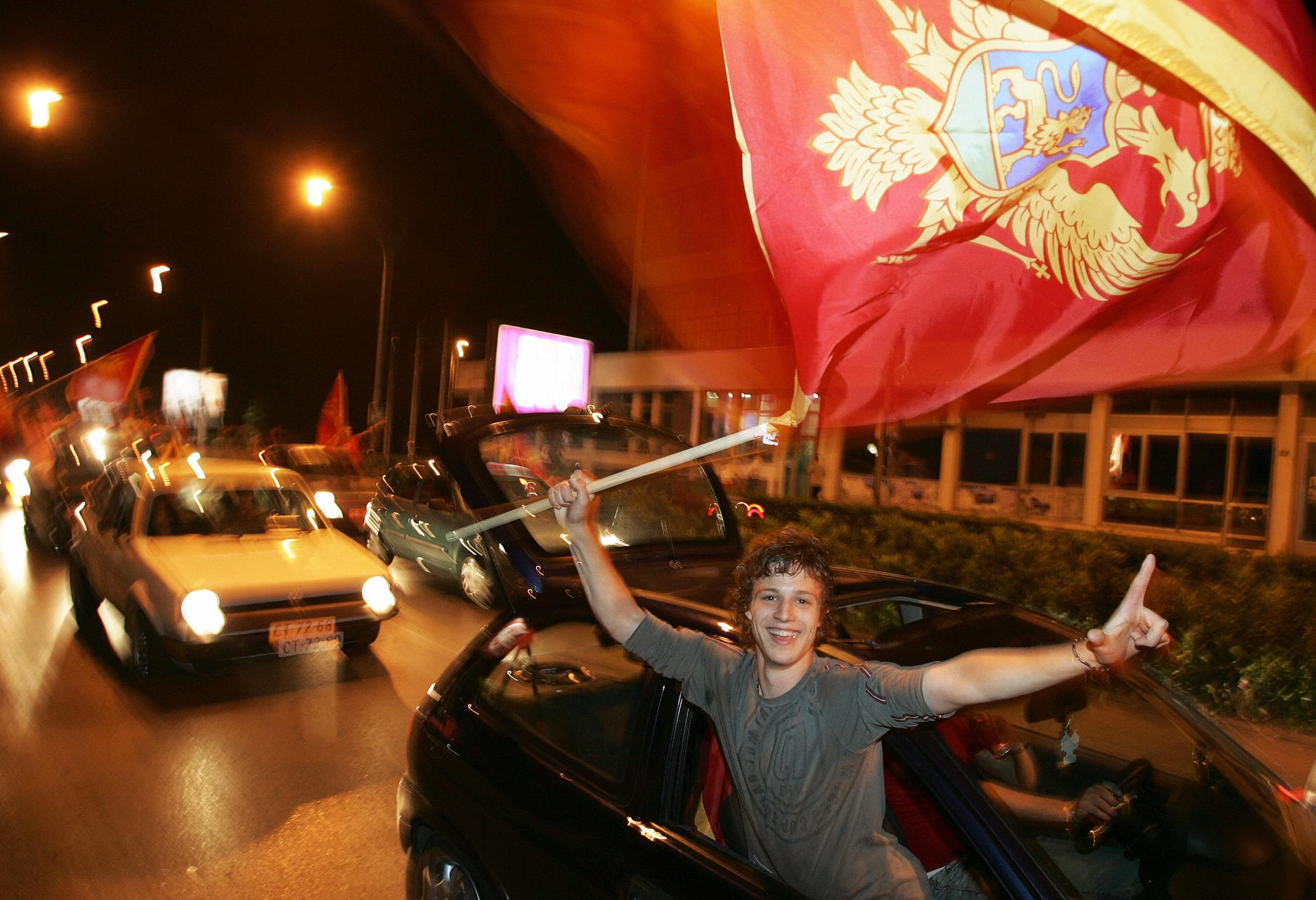 A man holds a flag and sticks his arm out of a car window, smiling and celebrating. 